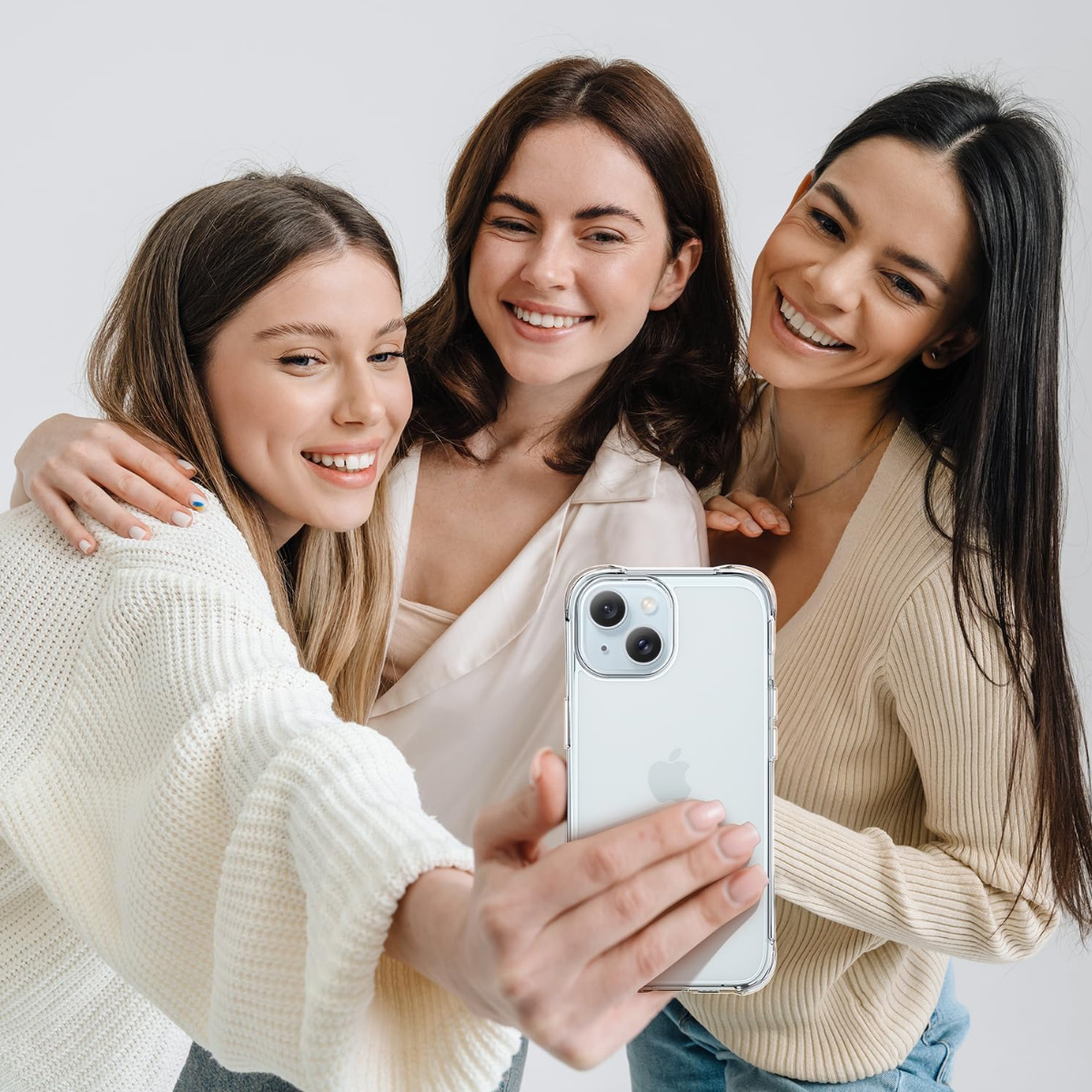 Drei lächelnde Frauen machen ein Selfie mit einem Handy. Eine hält das Telefon, andere haben sich gegenseitig in den Arm genommen, alle in hellen Kleidern.
