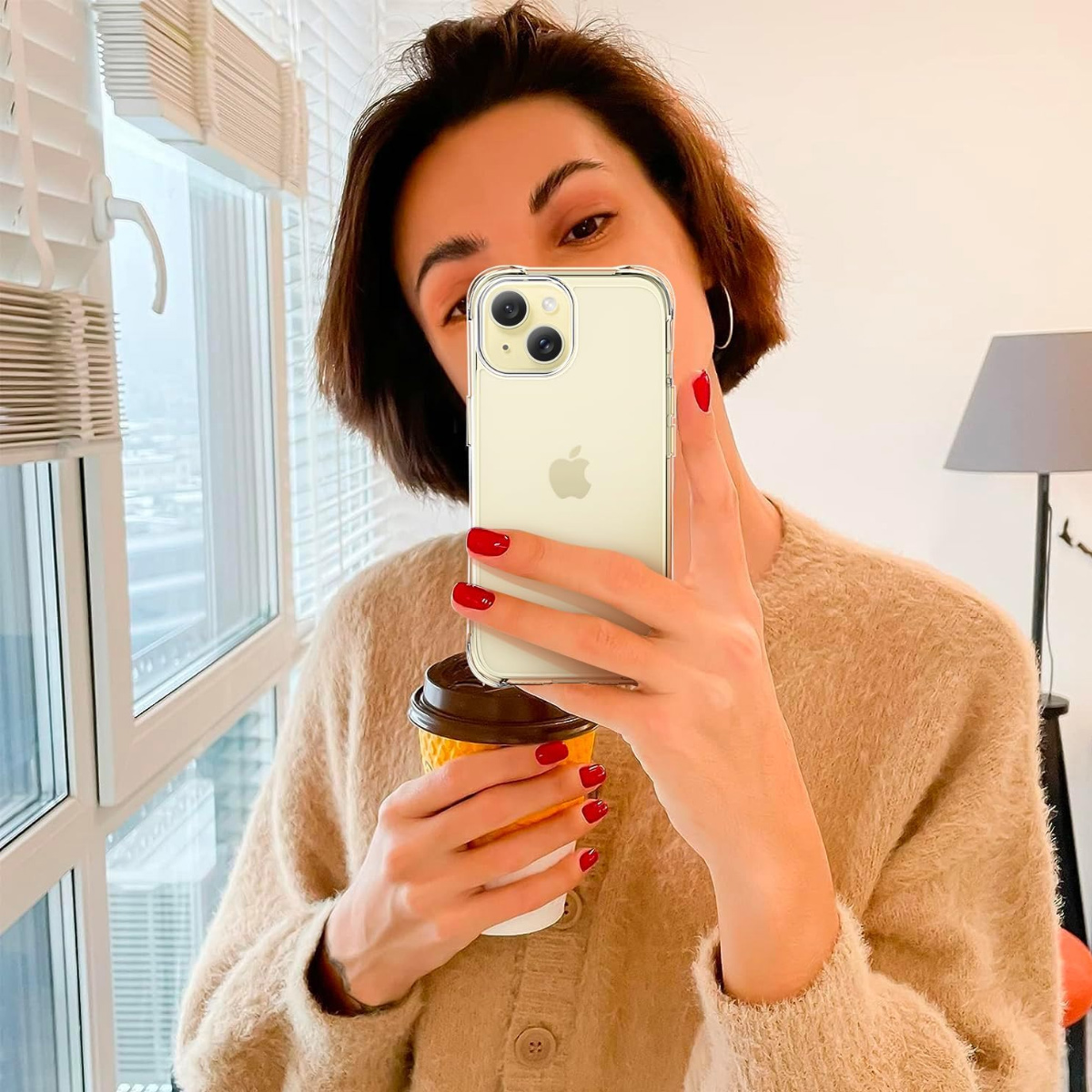 Frau hält Handy, Kaffeetasse. Rote Nägel, kurze Haare, beiger Pullover. Fenster, Lampe im Hintergrund. Macht ein Selfie.