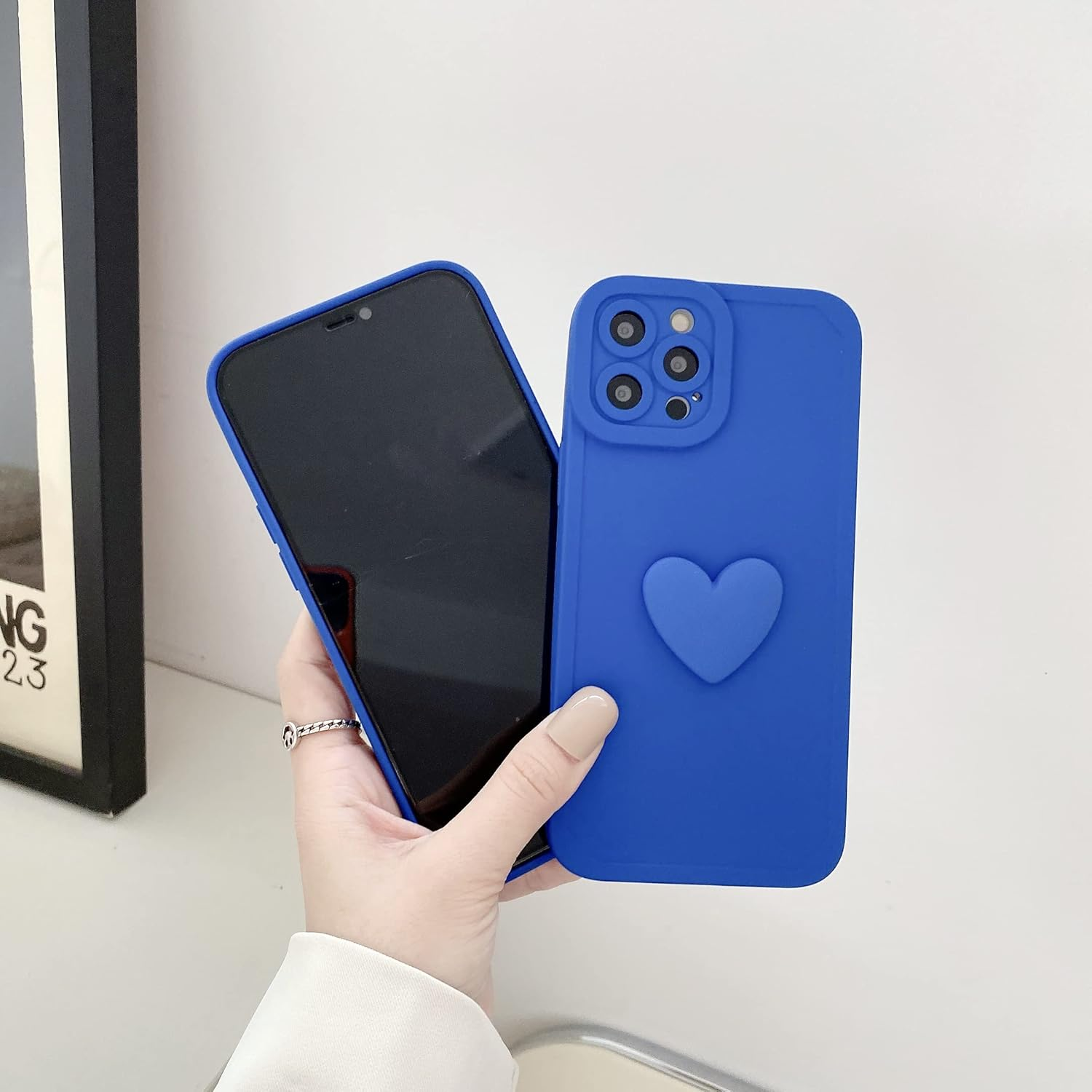 Eine Hand hält ein Telefon und eine blaue Handyhülle mit Herz. Das Telefon ist schwarz. Ein Bilderrahmen ist im Hintergrund.