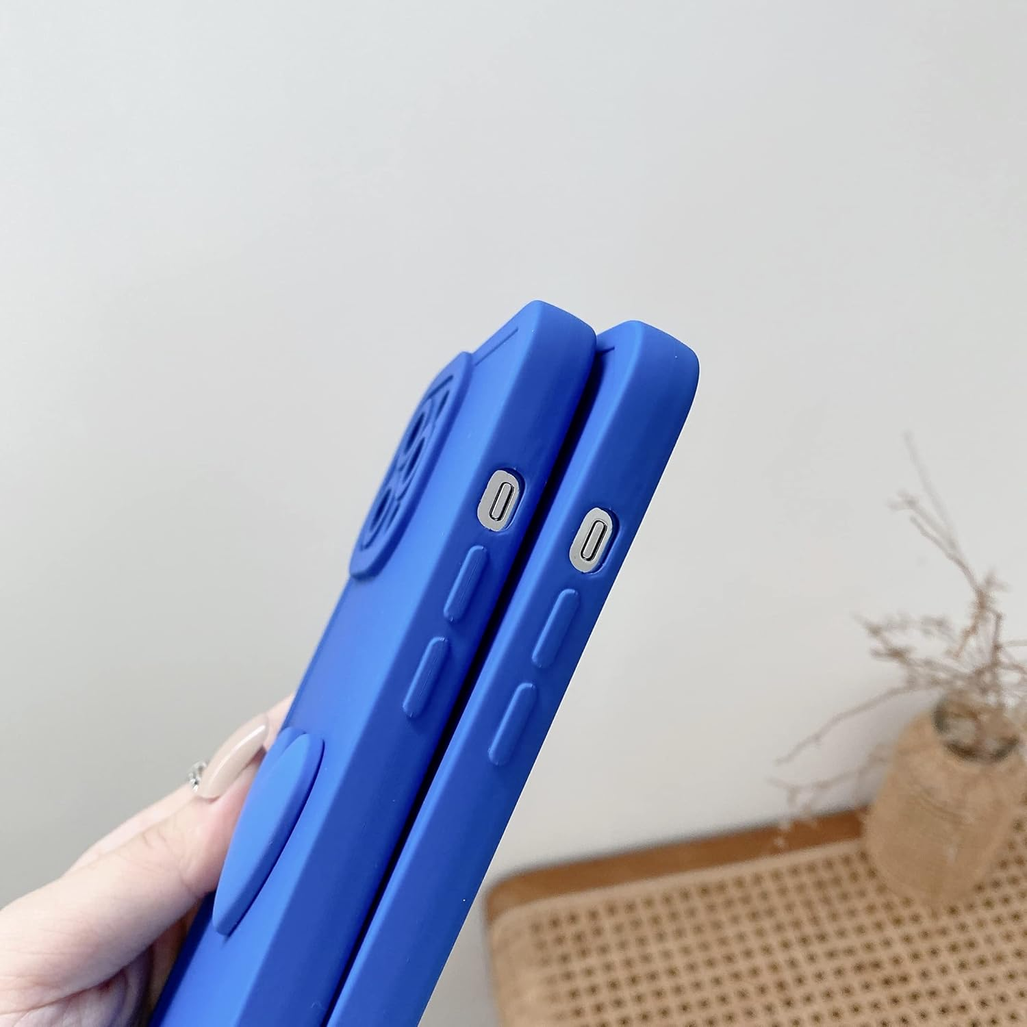 Zwei blaue Handyhüllen liegen nebeneinander. Eine Hand hält die Hülle. Der Hintergrund ist eine weiße Wand und ein Korbstuhl mit einer Pflanze.