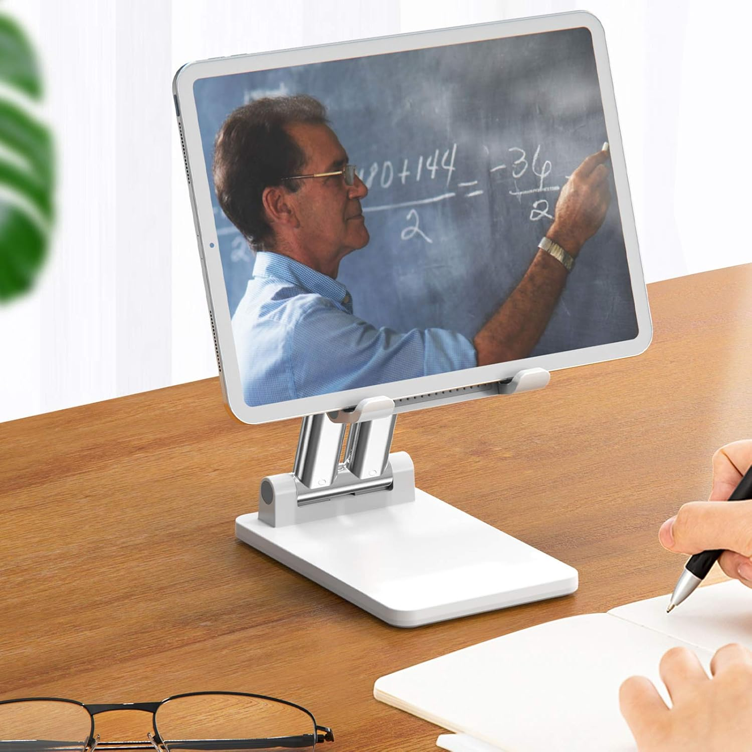 Tablet zeigt Mathelehrer, gehalten von weißem Ständer. Brille und Notizbuch auf Holzschreibtisch. Eine Hand schreibt.
