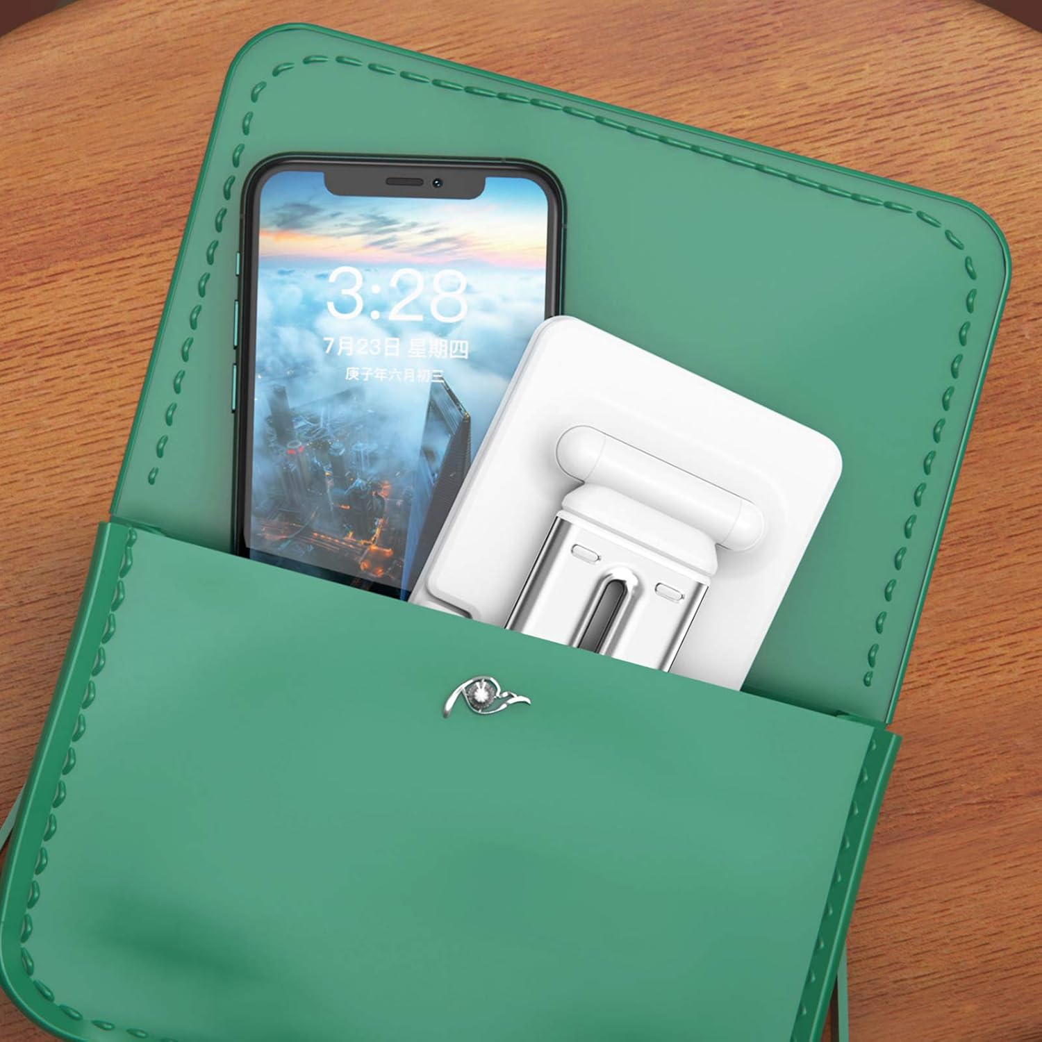 Eine grüne Handtasche enthält ein Smartphone, das die Uhrzeit anzeigt, und einen weißen Telefonständer. Die Tasche steht auf Holz.