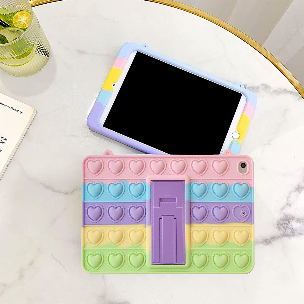 Ein iPad in einer mehrfarbigen Hülle mit Pop-It-Design, auf einem Marmortisch. Ein Getränk und ein Buch sind teilweise sichtbar.
