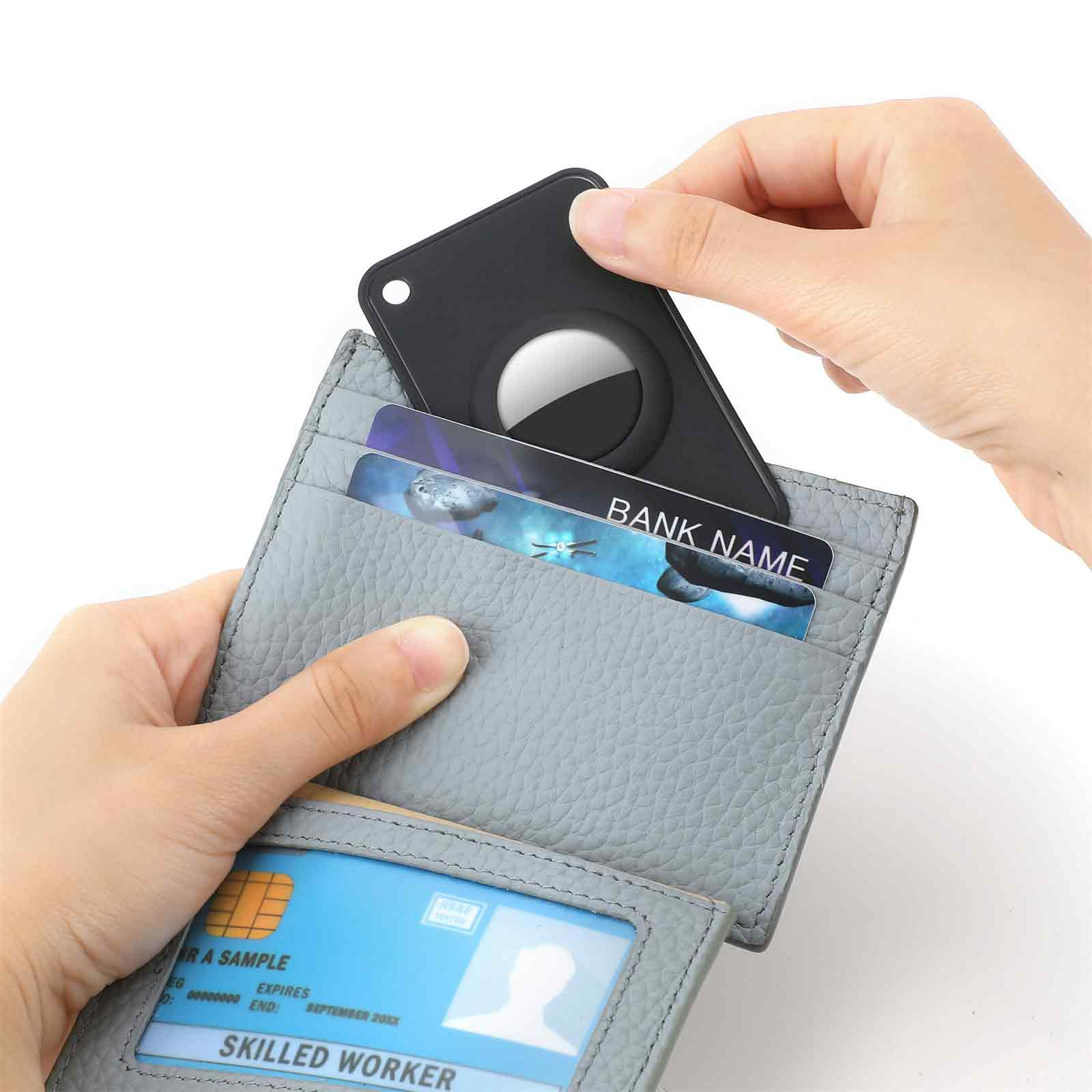 Eine Hand legt ein schwarzes AirTag in ein graues Portemonnaie mit Karten mit 'Bank Name' und Ausweisen. Der Hintergrund ist weiß.