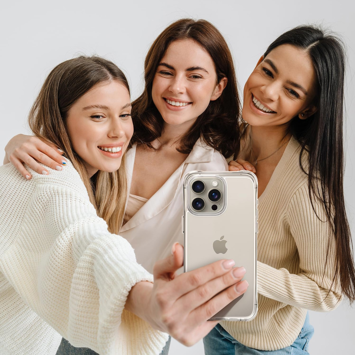 Drei lächelnde Frauen machen ein Selfie mit einem Telefon. Sie umarmen sich und tragen Freizeitkleidung. Weißer Hintergrund.