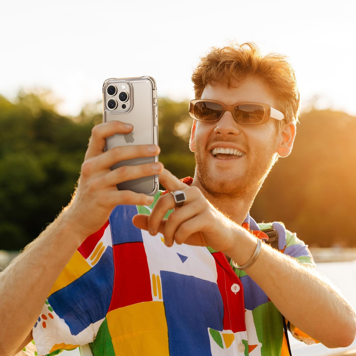 Ein Mann mit Sonnenbrille lächelt, während er ein Telefon hält. Er trägt ein buntes Hemd und einen Ring. Der Hintergrund zeigt Bäume und Sonnenlicht.