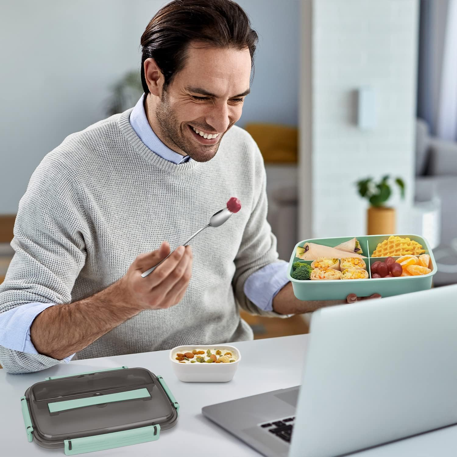 Mann lächelt, isst mit einem Löffel. Lunchbox mit Essen in der Hand. Laptop auf dem Tisch. Eine weitere Lunchbox und eine Schüssel mit Nüssen.