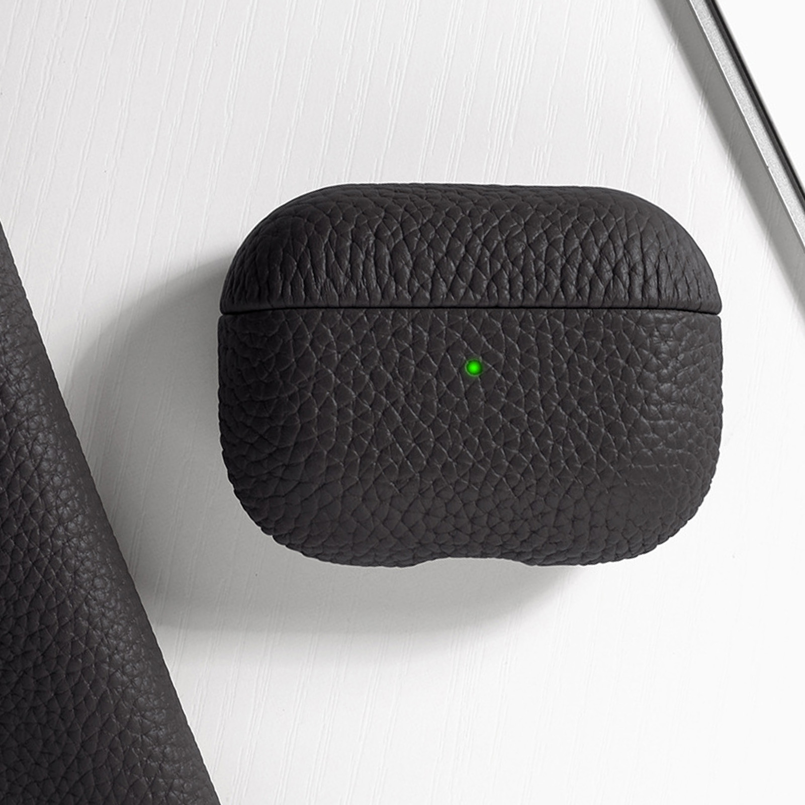 Schwarzes Leder-AirPods-Etui mit grünem Licht auf weißer Oberfläche. Ein Stück Leder ist links.