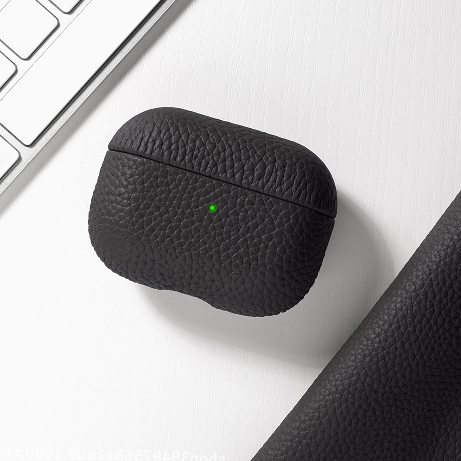 Schwarzes, strukturiertes AirPods-Etui auf weißer Oberfläche, neben Tastatur und dunklem Lederimitat.