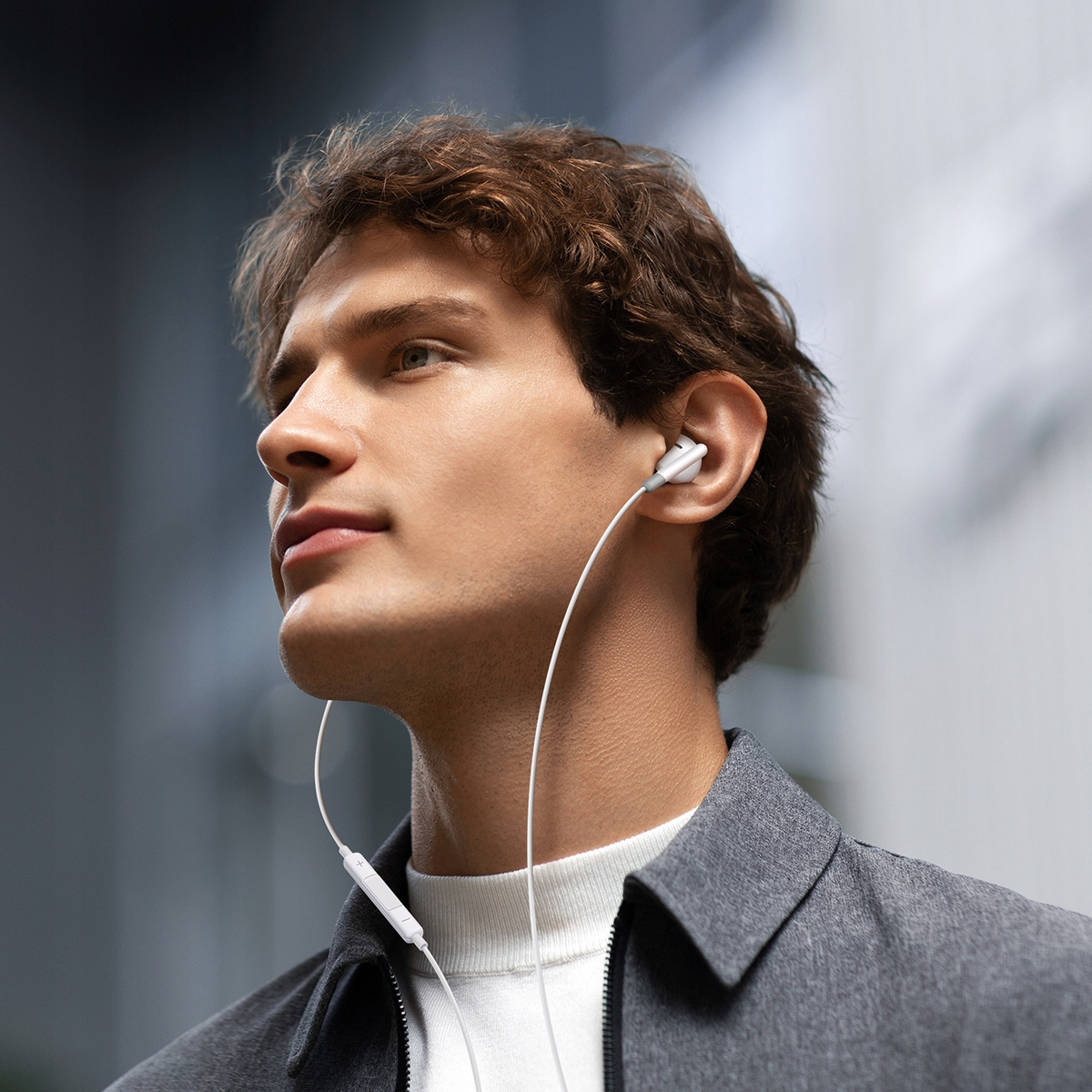 Mężczyzna w słuchawkach patrzy w bok. Ma brązowe włosy i szarą kurtkę. Tło jest rozmyte.