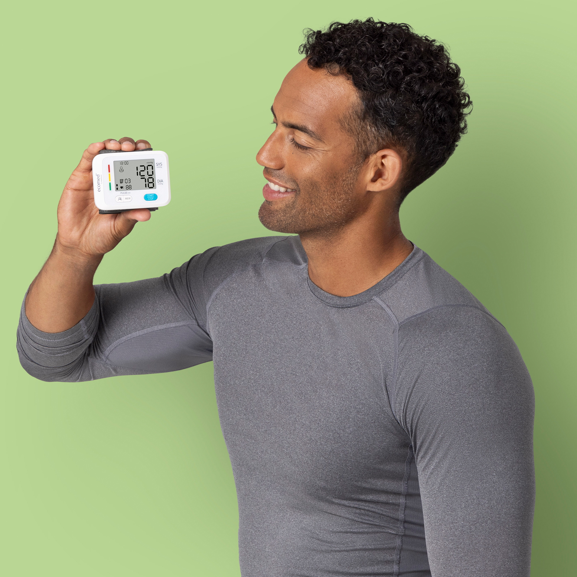 Mężczyzna trzymający monitor ciśnienia krwi, uśmiechnięty. Monitor wyświetla liczby. Ma na sobie szarą koszulę. Zielone tło.