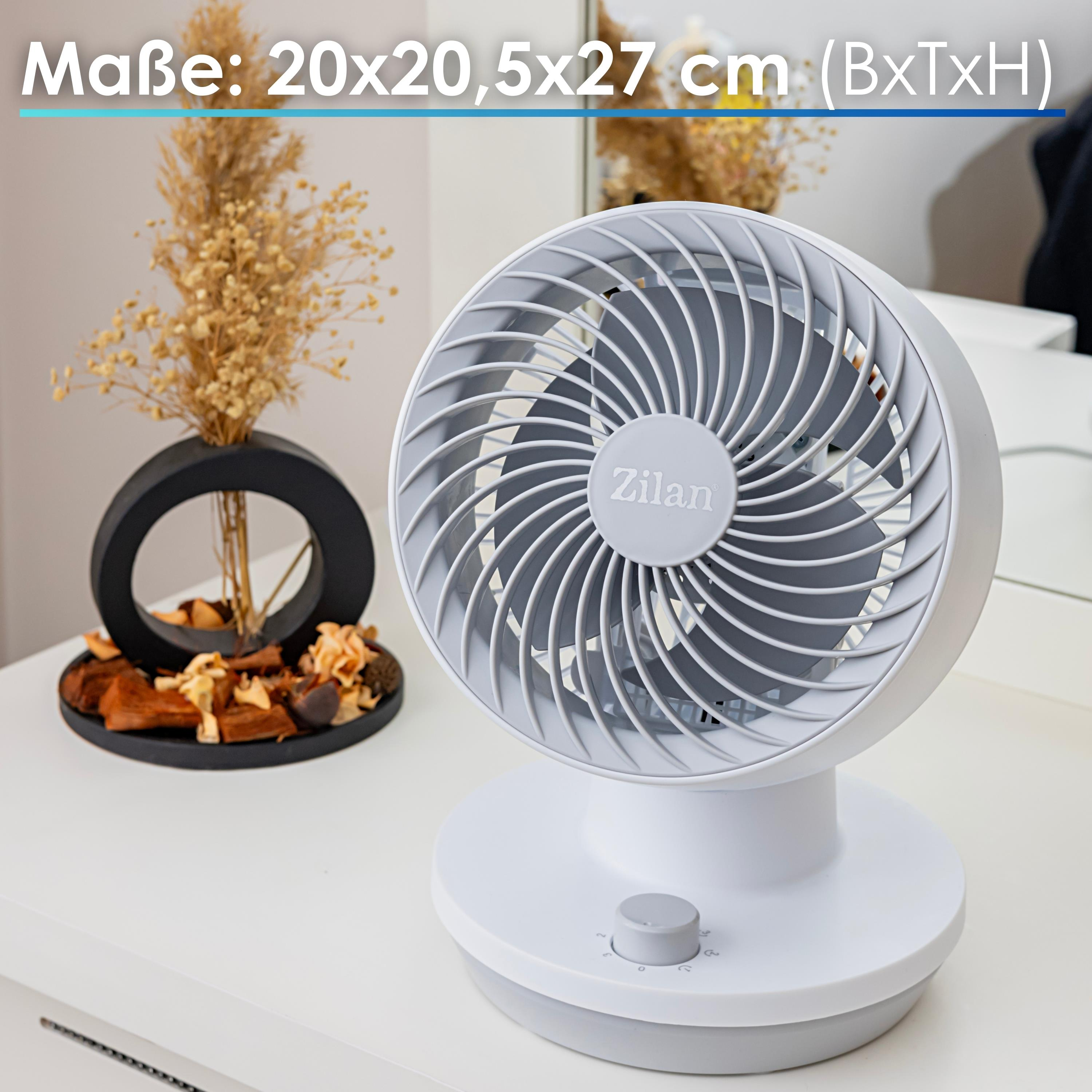Ein weißer Ventilator mit grauen Blättern und einem Drehregler steht auf einem weißen Tisch. Links eine schwarze Vase mit Trockenblumen.