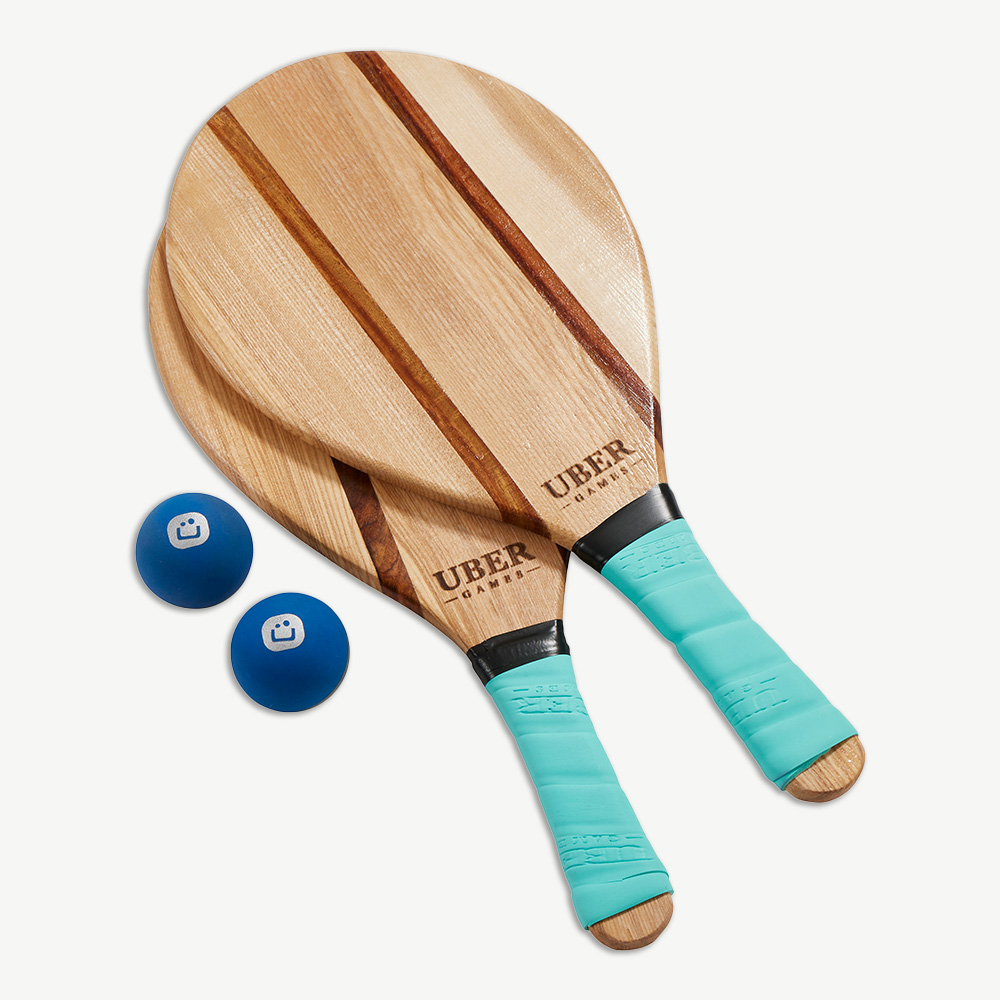 Zwei Holz-Beach-Tennis-Schläger und zwei blaue Bälle mit weißem Smiley-Gesicht-Logo auf weißem Untergrund.