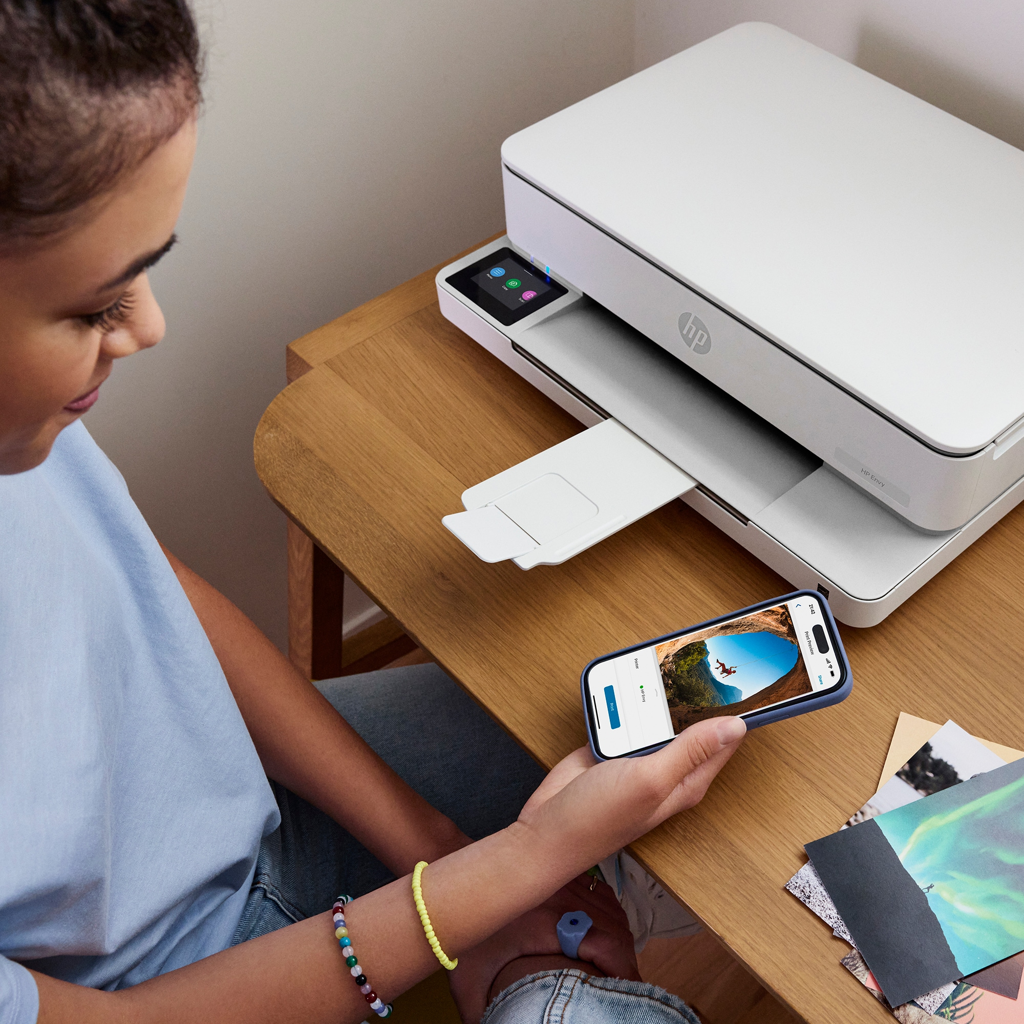 Een vrouw houdt een telefoon vast met een afbeelding, in de buurt van een witte printer op een houten bureau. Er liggen afdrukken op het bureau.
