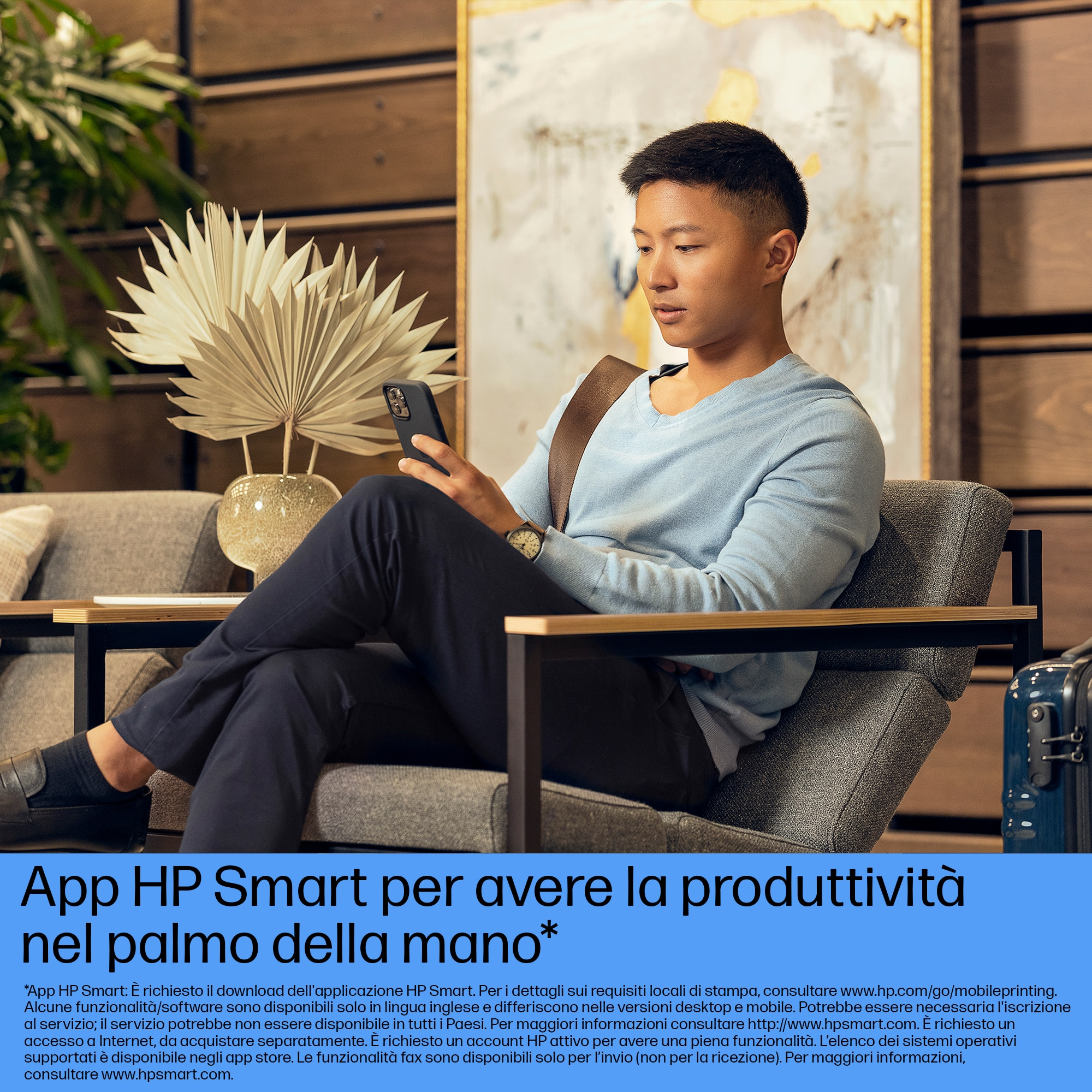 Mężczyzna siedzi i patrzy na telefon. Jest w nowoczesnej przestrzeni. W tle jest roślina i sztuka.