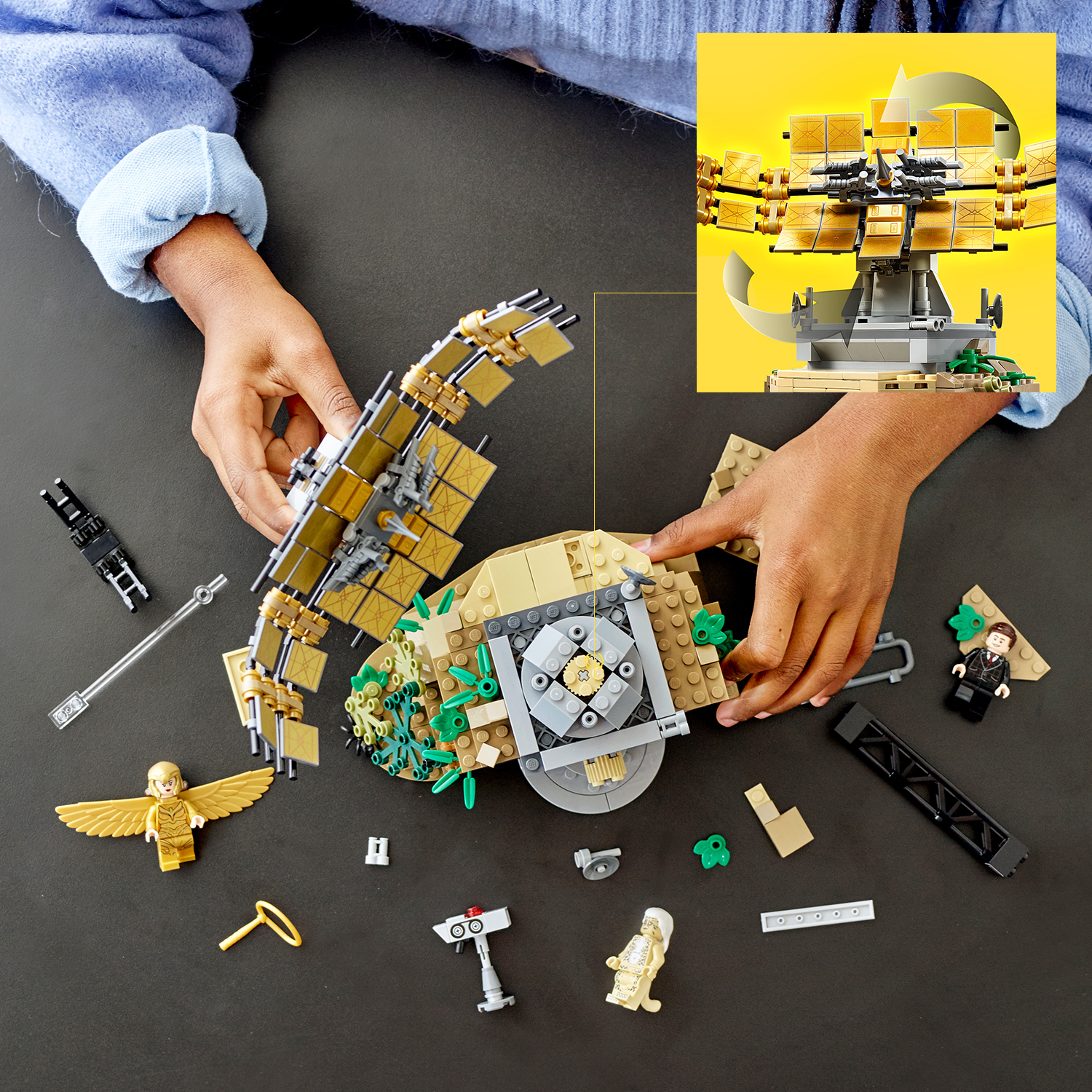 Hände bauen ein Lego-Set zusammen. Enthält Lego-Figuren, goldene und beige Bauteile und eine schwarze Oberfläche.