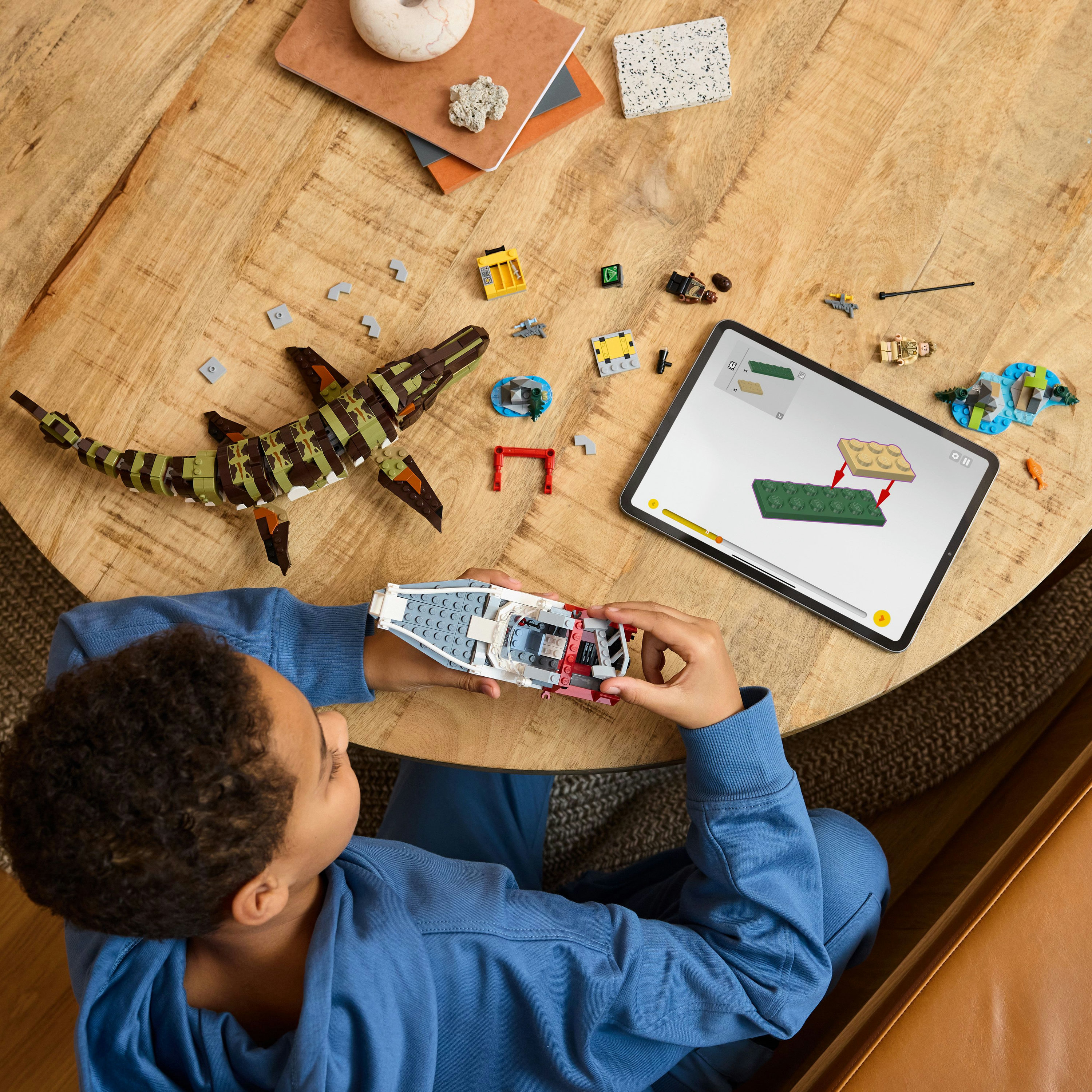 Dziecko buduje model Lego łodzi, z tabletem pokazującym instrukcje. Drewniany stół zawiera inne części Lego.