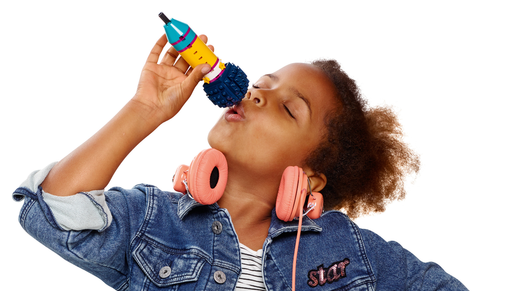 Dziewczyna z zamkniętymi oczami śpiewa do kolorowego mikrofonu. Ma na sobie dżinsową kurtkę i różowe słuchawki.