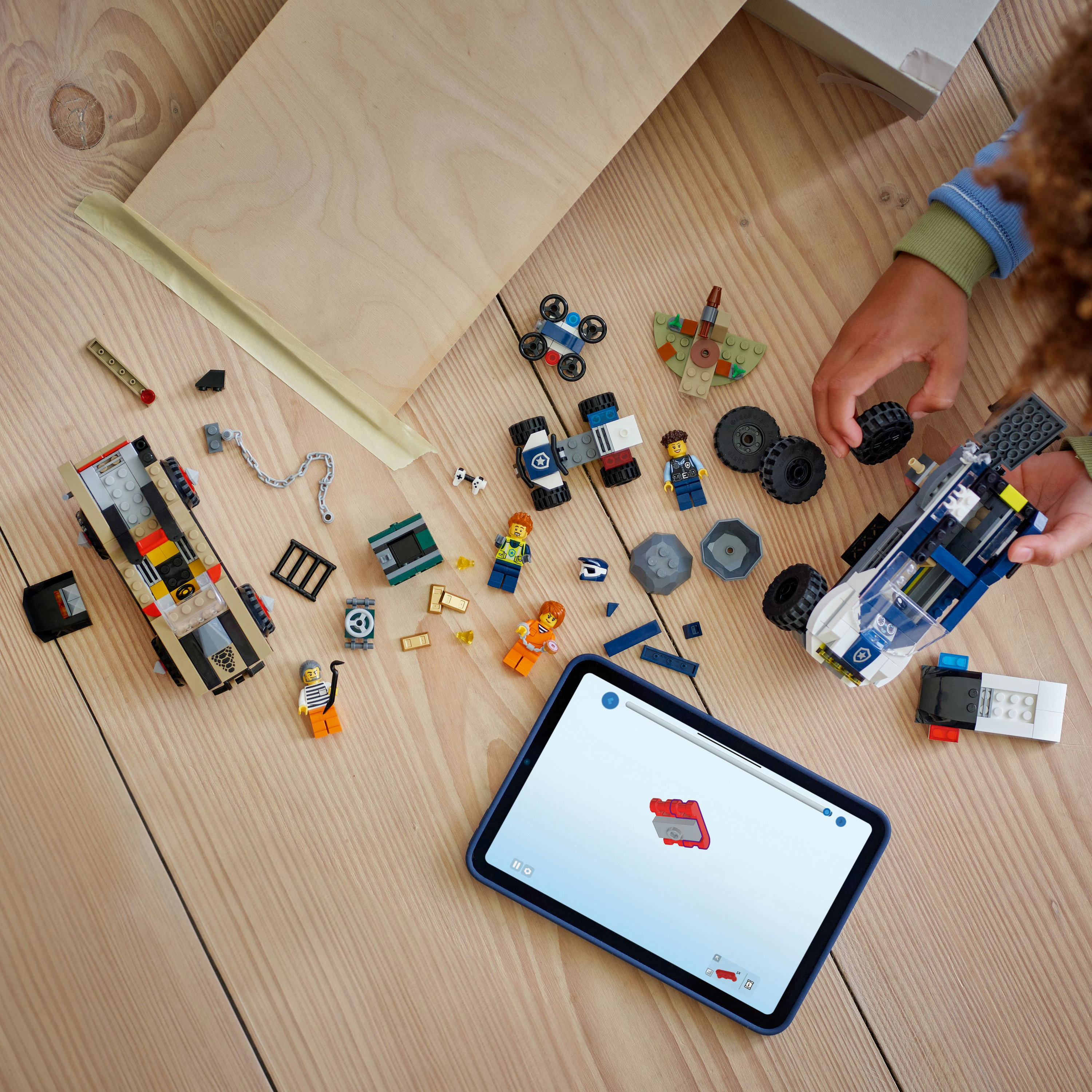 Dziecko buduje z klocków Lego na drewnianej powierzchni. Tablet wyświetla instrukcje. Różne zestawy Lego są rozrzucone.