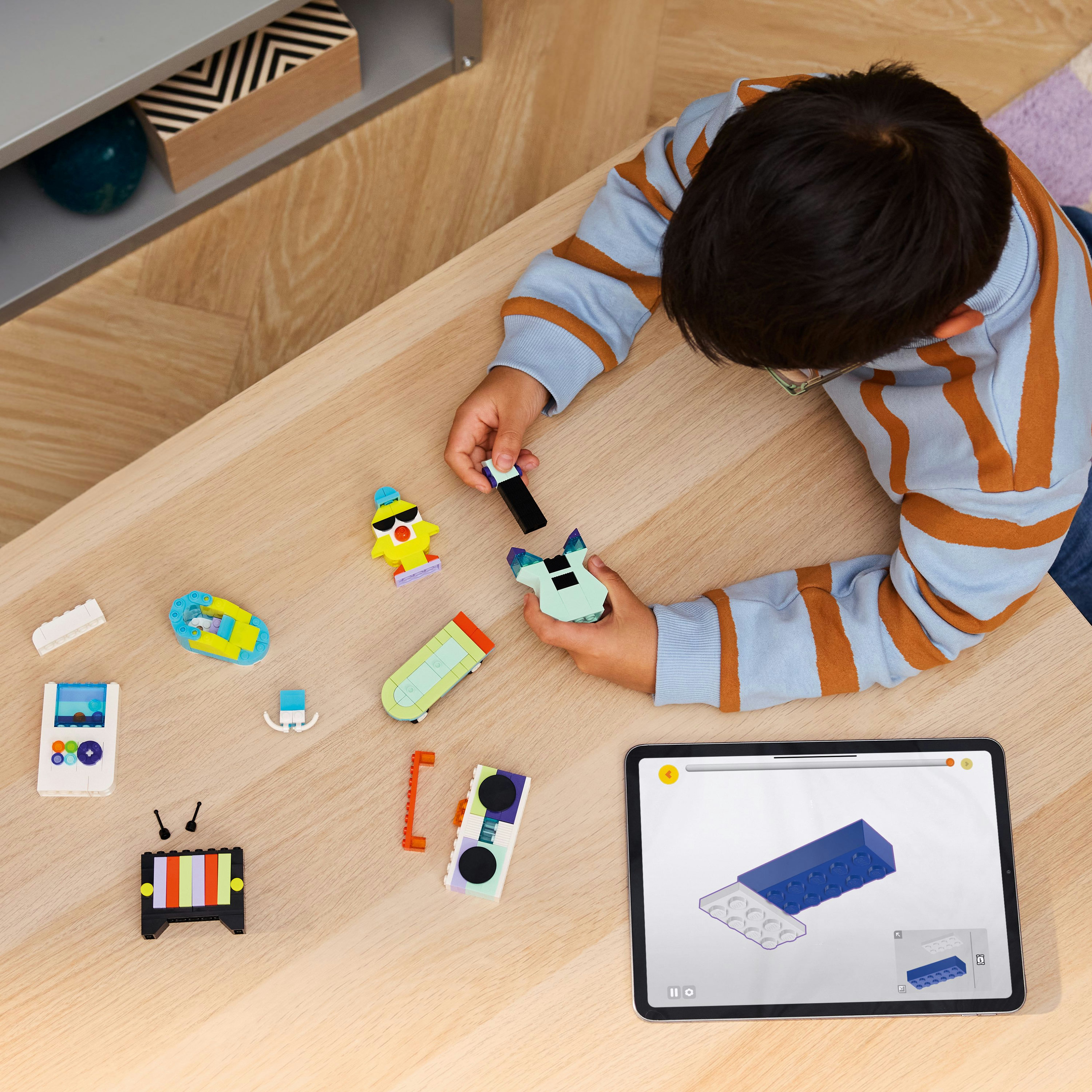 Bir çocuk, ahşap bir masada Lego oyuncaklarını birleştiriyor, talimatları gösteren bir tabletle. Oyuncaklar etrafa dağılmış.