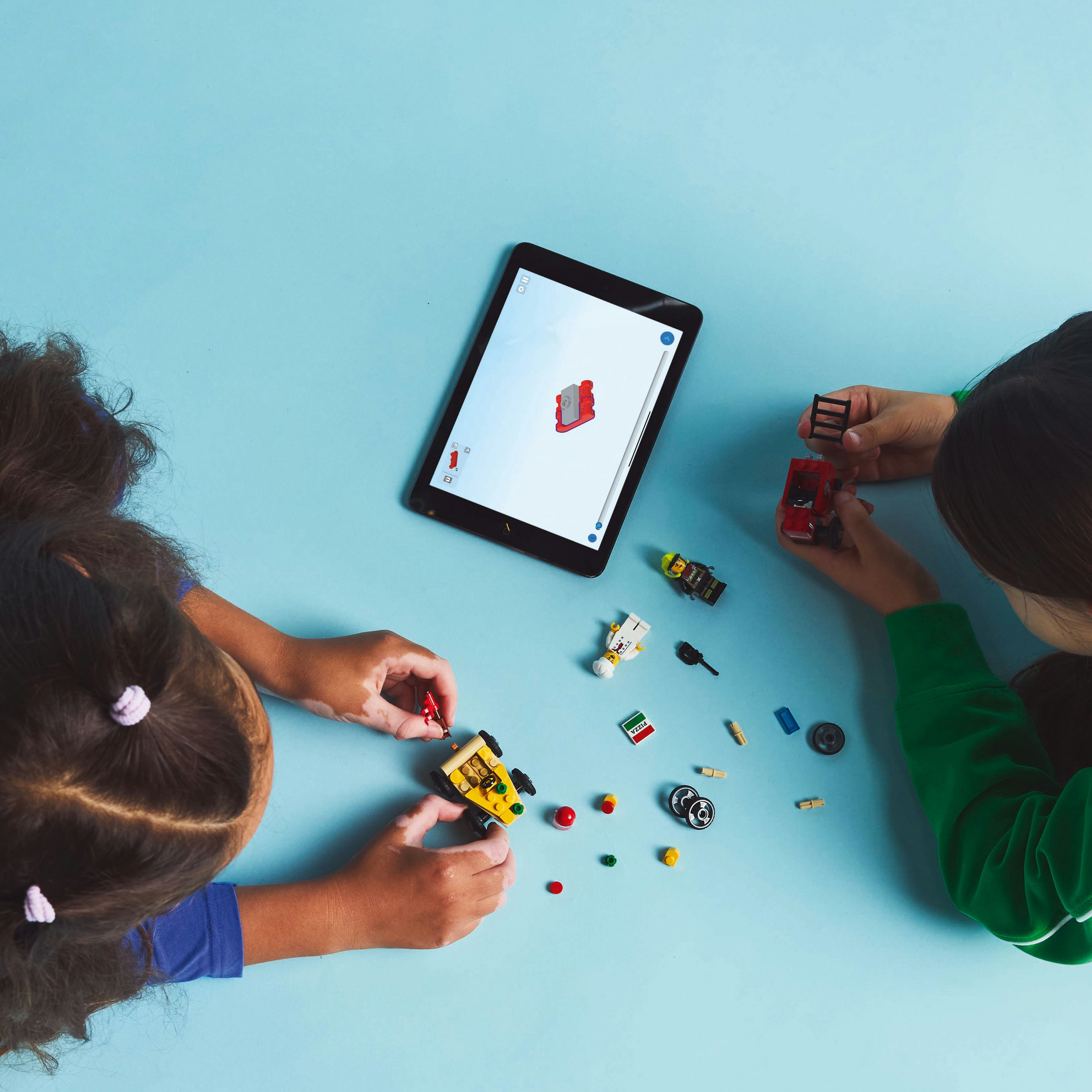 Dos niños construyen con Lego sobre una superficie azul. Una tableta muestra instrucciones. Piezas y figuras de Lego dispersas.