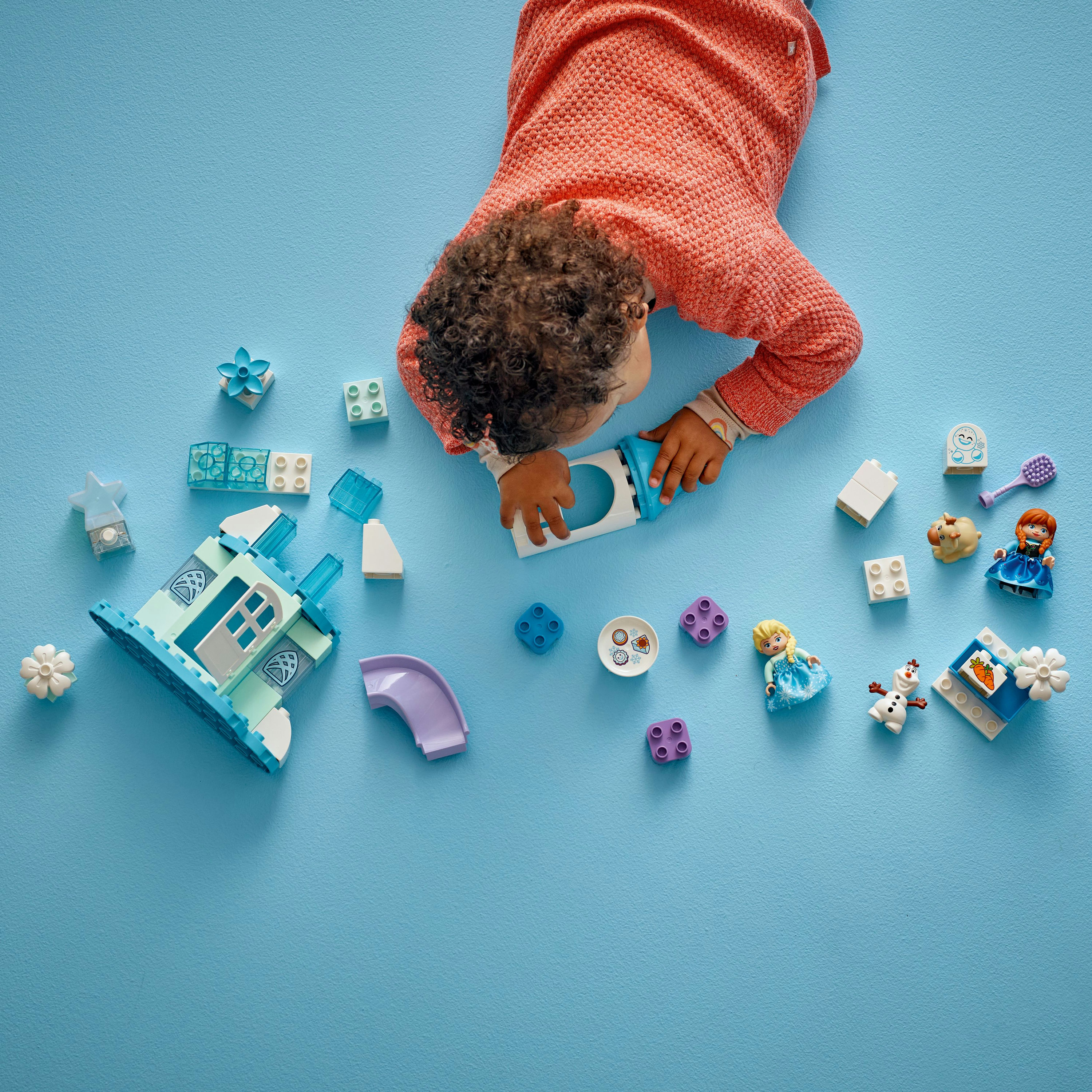 Dziecko leży na niebieskiej powierzchni z klockami i figurkami. Dziecko ma na sobie pomarańczowy sweter.