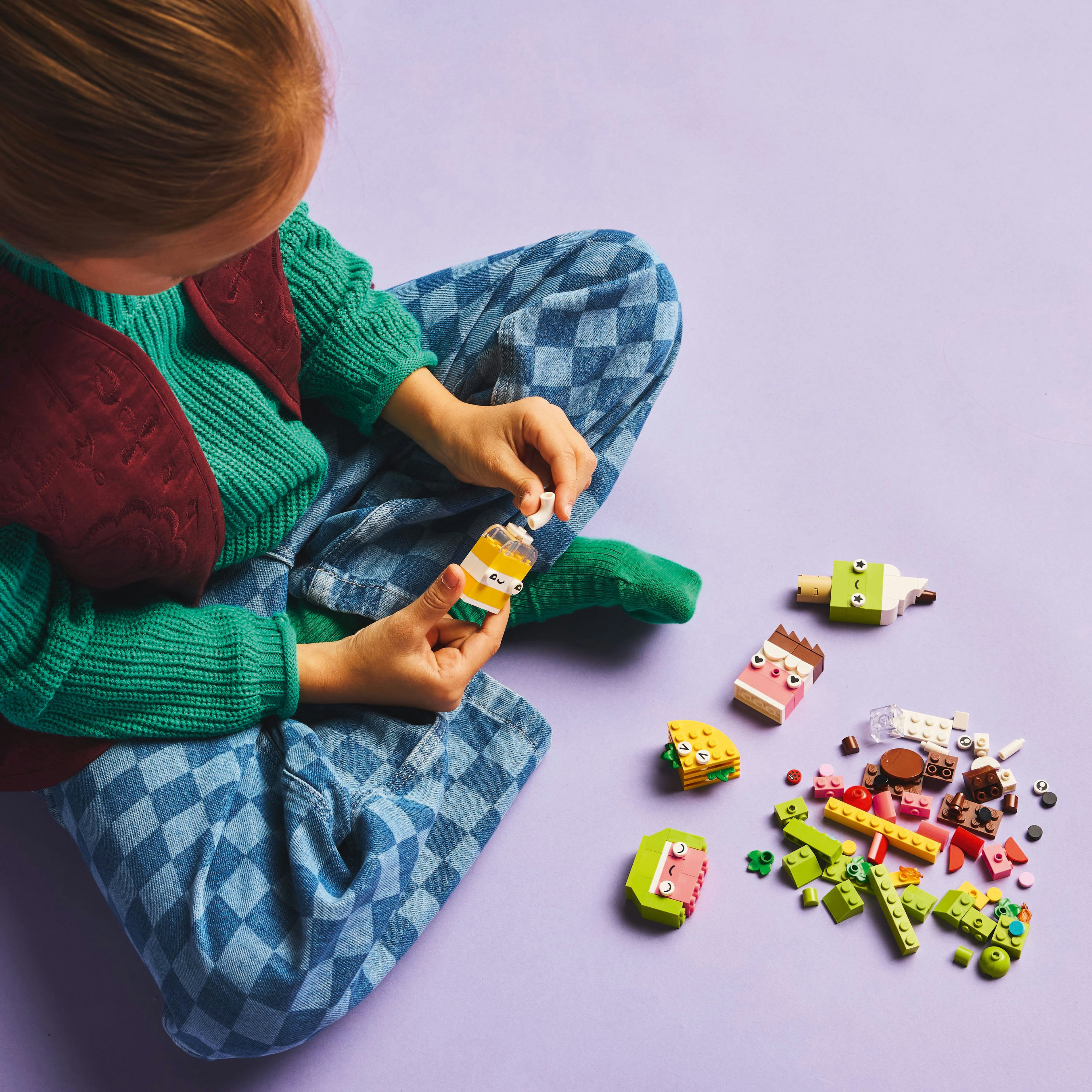 Dziecko siedzi, składając zabawkowe jedzenie. Kawałki jedzenia Lego są rozrzucone na fioletowej powierzchni, w tym napój, lody i burger.