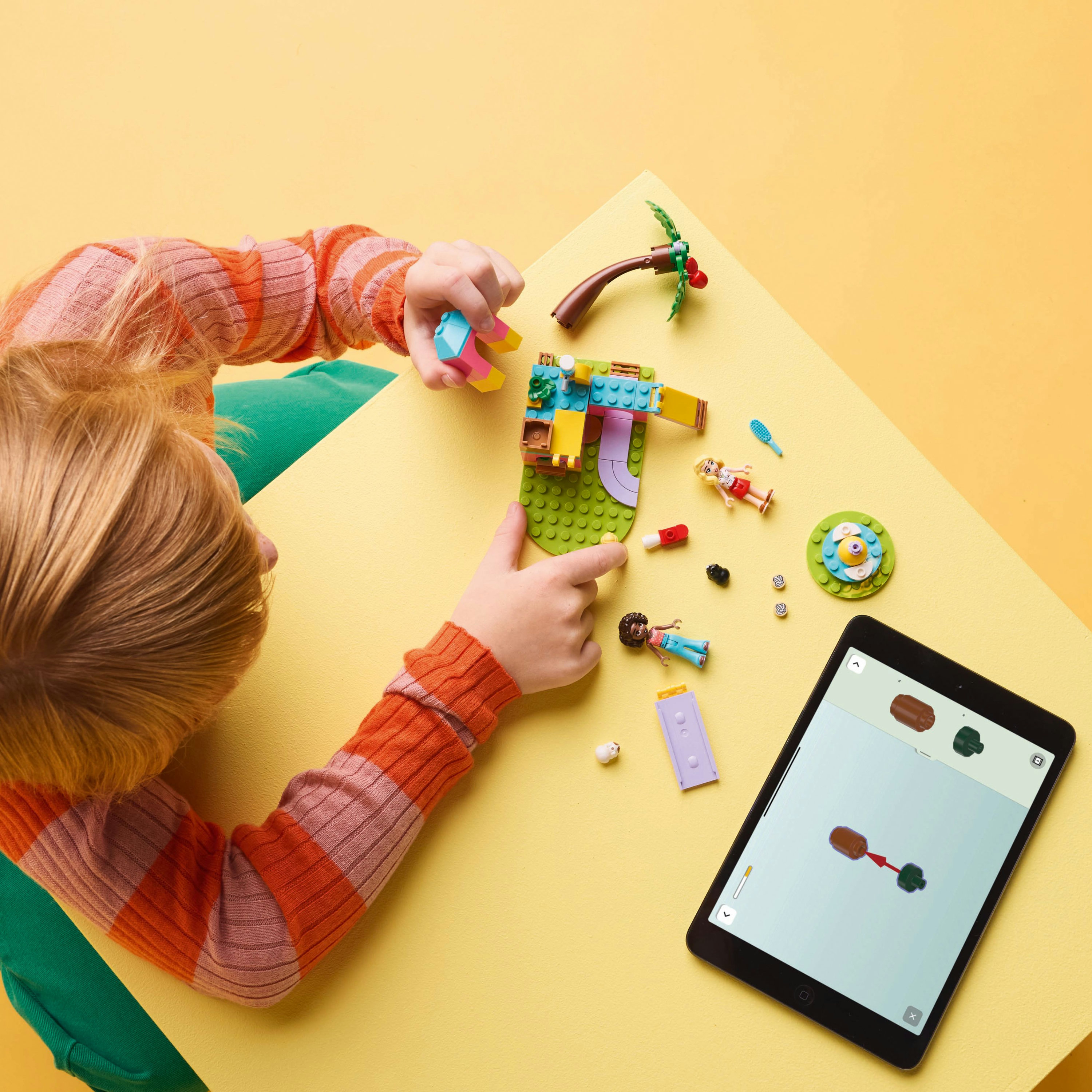 Un bambino sta assemblando un set Lego, con un iPad che mostra le istruzioni. Vari pezzi e personaggi Lego sono sparsi.