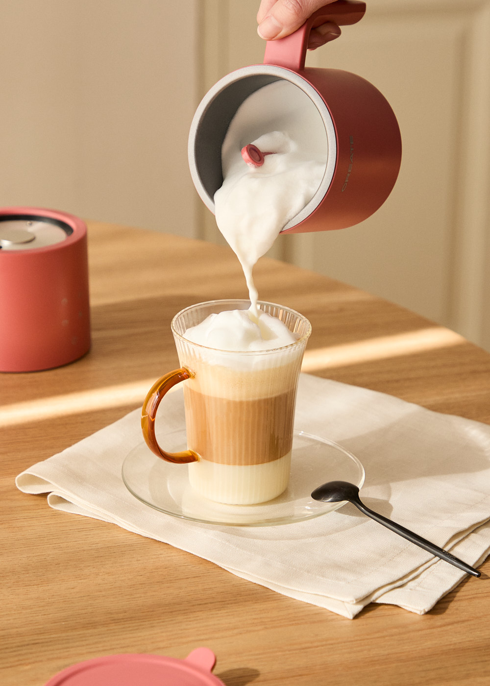 Una persona vierte espuma de leche de un espumador rosa en una bebida de café en capas. La bebida está en un platillo con una cuchara.