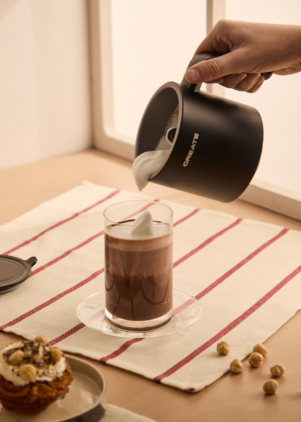Una mano vierte espuma de una jarra negra en un vaso de chocolate. Pasteles y nueces están en la mesa.