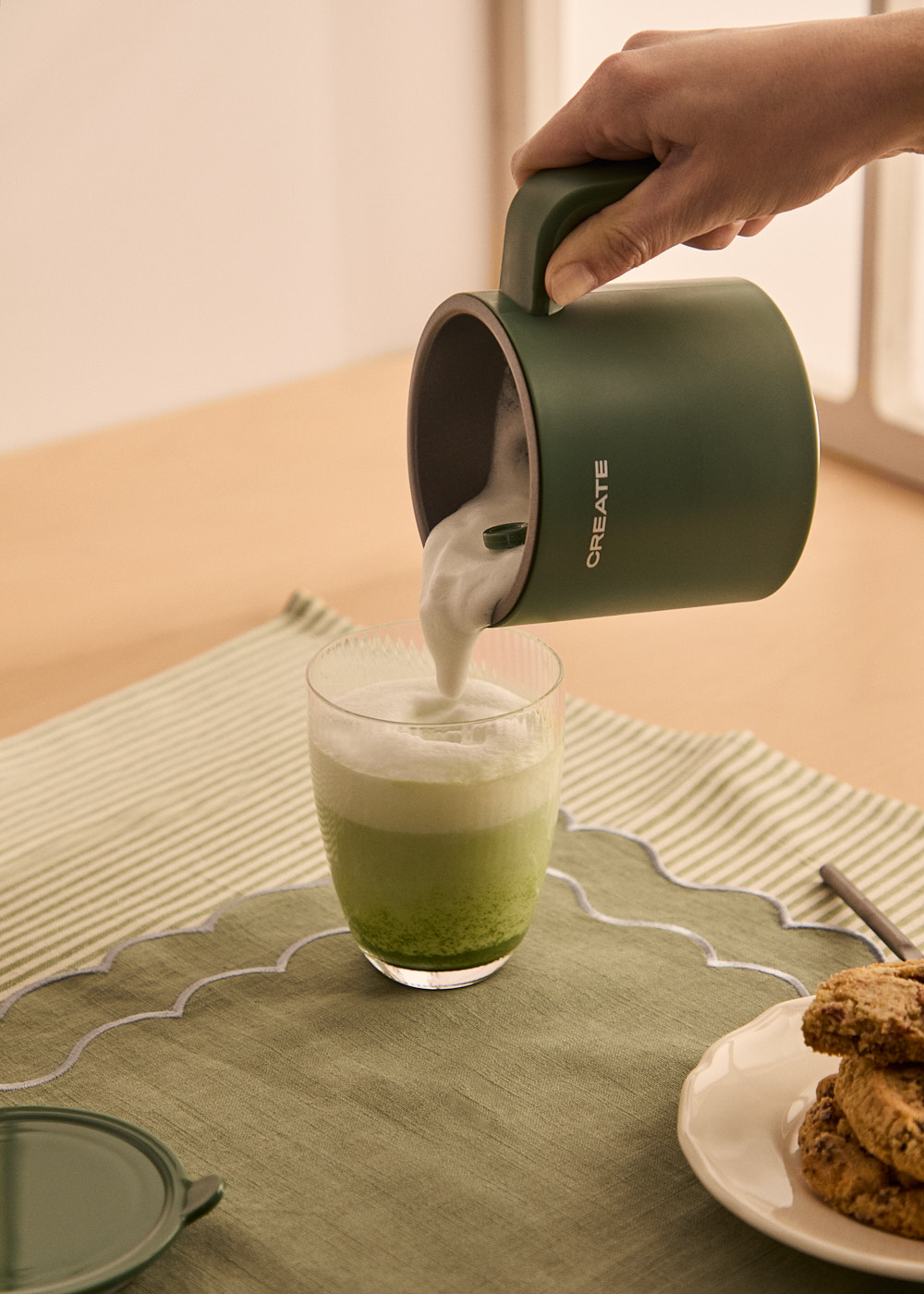 Una mano vierte espuma en un vaso de matcha. Galletas en un plato y una tapa verde sobre una mesa con un mantel verde.