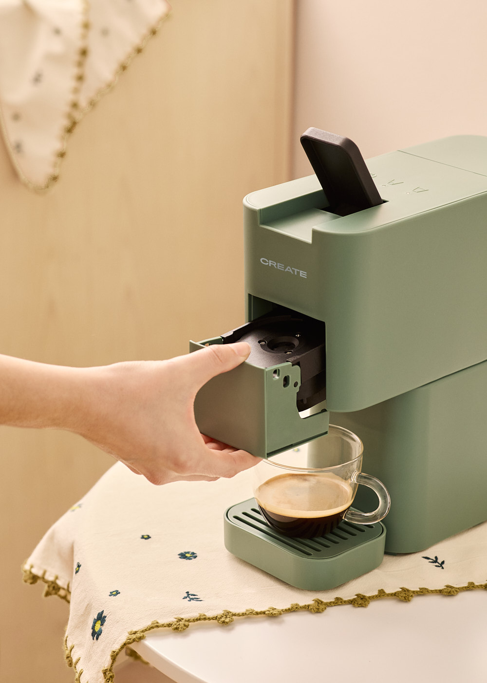 Una mano saca una bandeja de una cafetera verde. Una taza de café sobre un posavasos. Se ve un paño floral.