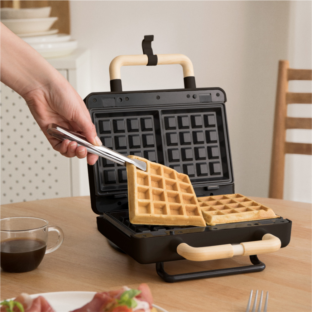 Una mano usa pinzas para sacar un gofre de una gofrera. Una taza de café está en una mesa junto a la plancha.