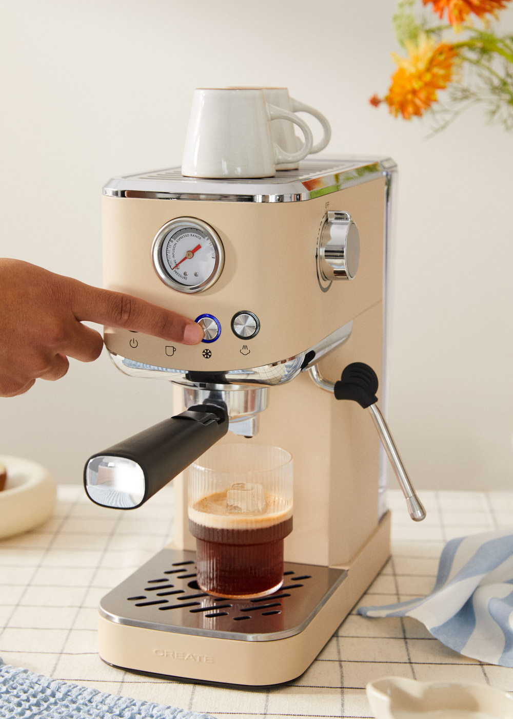 Un dedo presiona el botón de la máquina de espresso. Una taza de café está en la máquina. Dos tazas están encima.