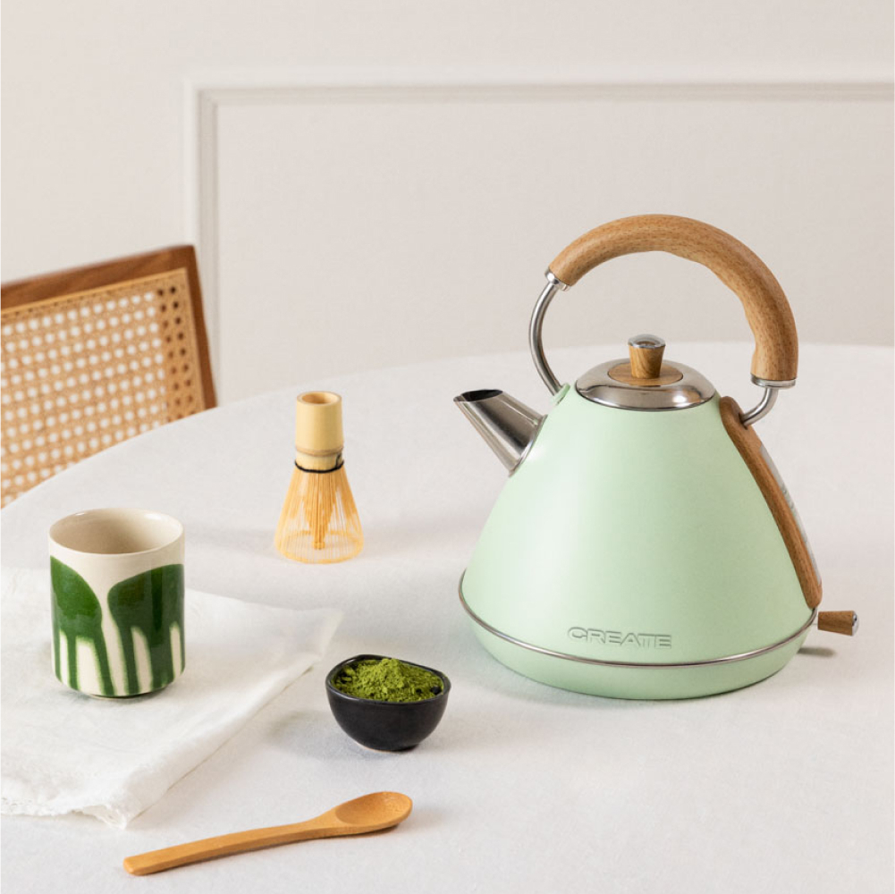 Ein mintgrüner Wasserkocher, eine Matcha-Schale, ein Schneebesen und eine Teetasse auf einem weißen Tisch. Holzlöffel und Stuhl im Hintergrund.
