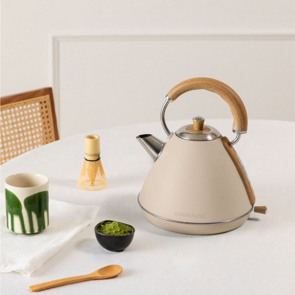 Una tetera crema con mango de madera sobre mesa blanca. Té matcha, batidor y taza en la mesa. Una silla en el fondo.