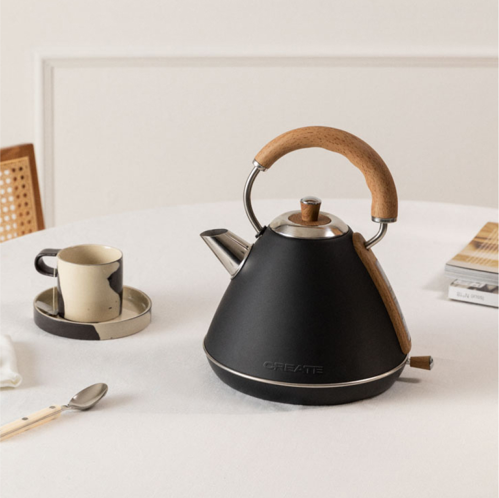 Ein schwarzer Wasserkocher mit Holzgriff und eine Tasse auf einer Untertasse stehen auf einem weißen Tisch. Ein Löffel und eine Zeitschrift sind ebenfalls zu sehen.