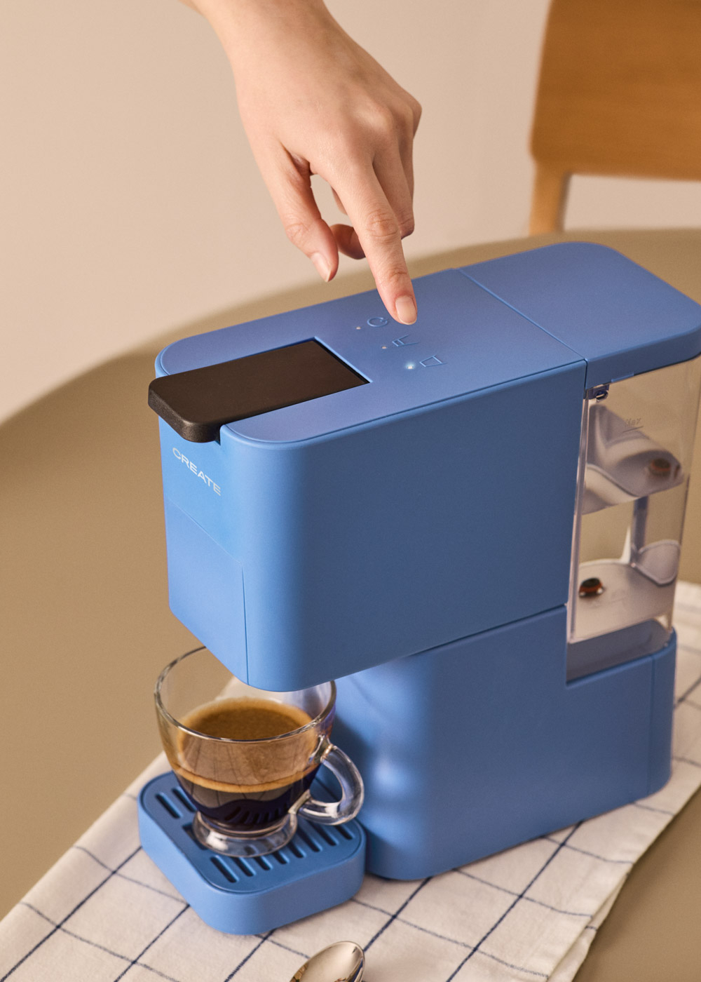 Un dedo presiona un botón en una cafetera azul. Una taza de vidrio con café está debajo. El fondo es neutro.