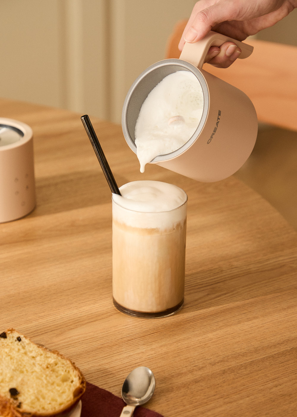 Una persona vierte leche espumada en un vaso con café. Una pajita negra está en el vaso. Una rebanada de pan y una cuchara están en la mesa.