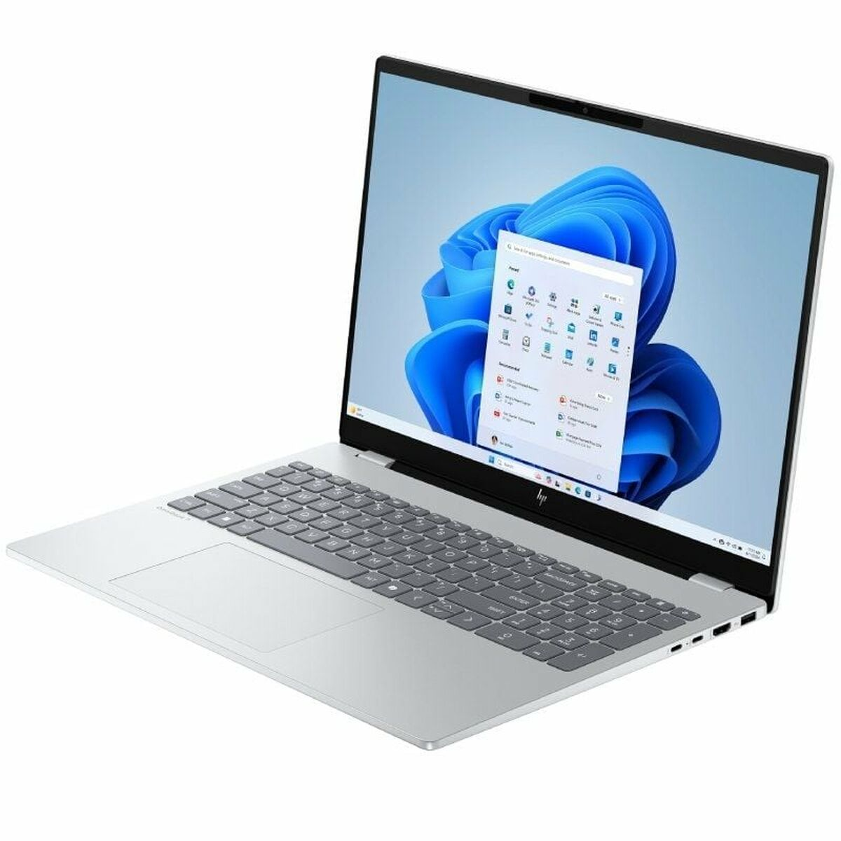 Laptop plateado muestra una pantalla de Windows 11 con fondo azul. El teclado es gris y la pantalla muestra iconos de aplicaciones.