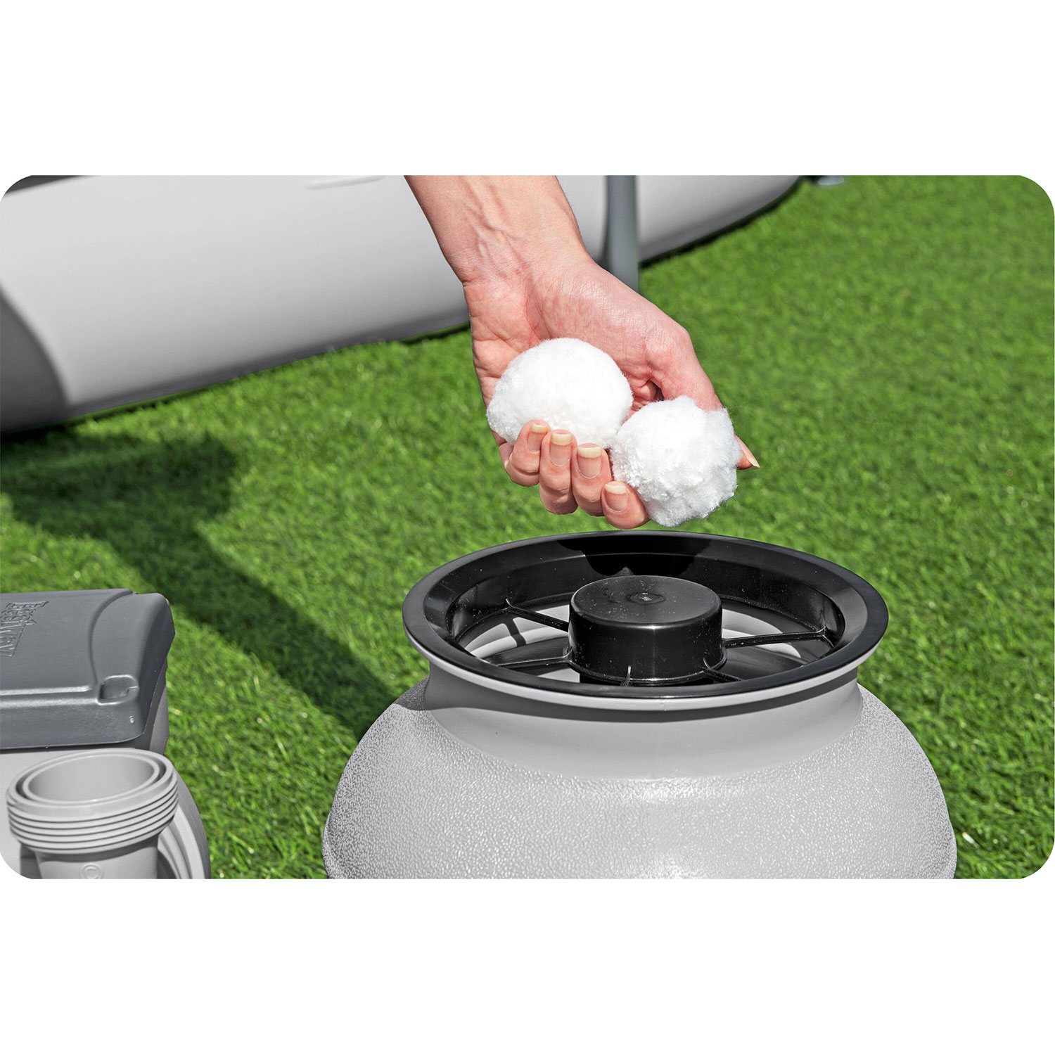 Ręka trzyma dwie białe kule filtrujące nad szarym filtrem. Zielona trawa w tle i szary obiekt po lewej.