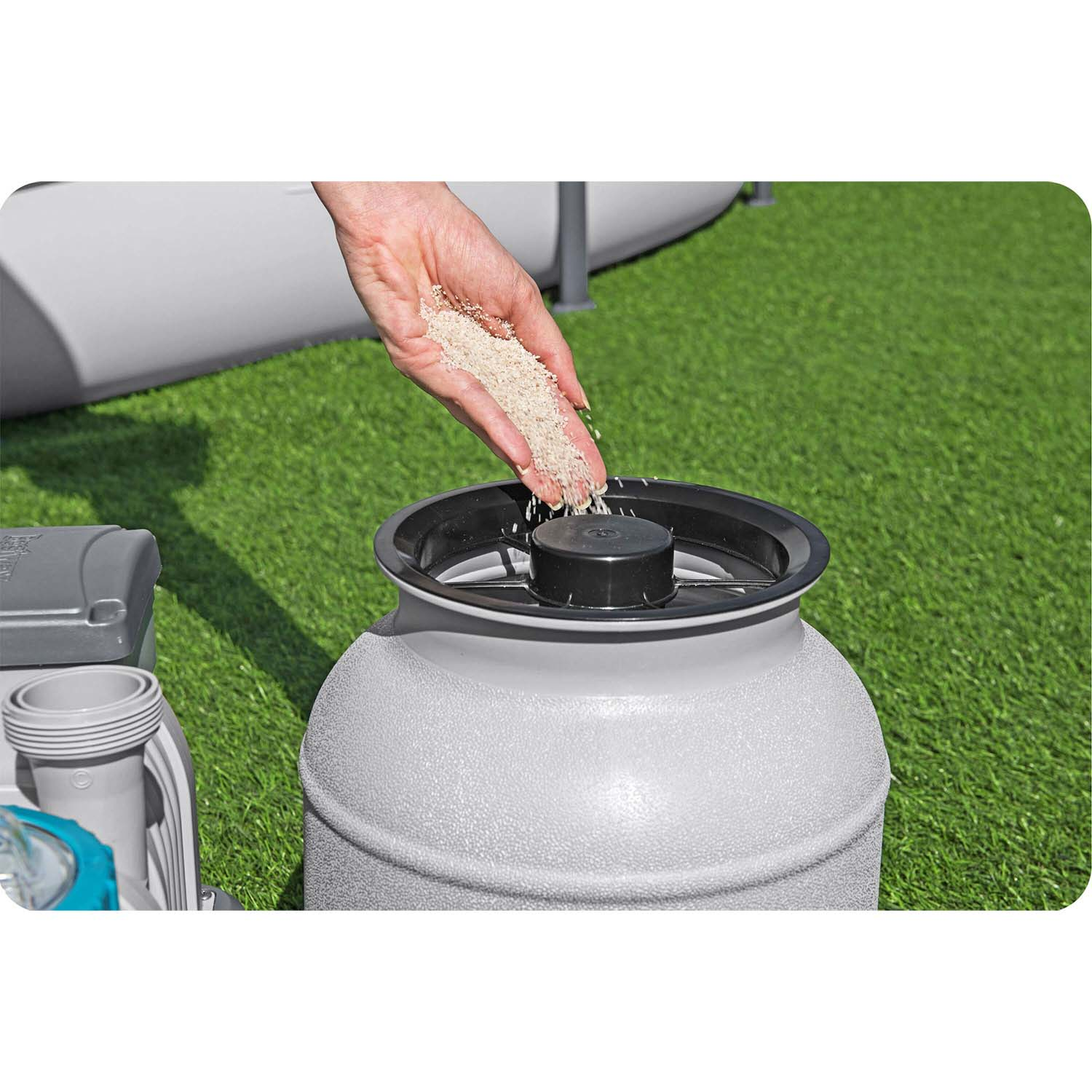 Ręka wsypuje piaskopodobny materiał do szarego pojemnika. Pojemnik ma czarną pokrywę i stoi na zielonej trawie. Po lewej znajduje się inny szary obiekt.