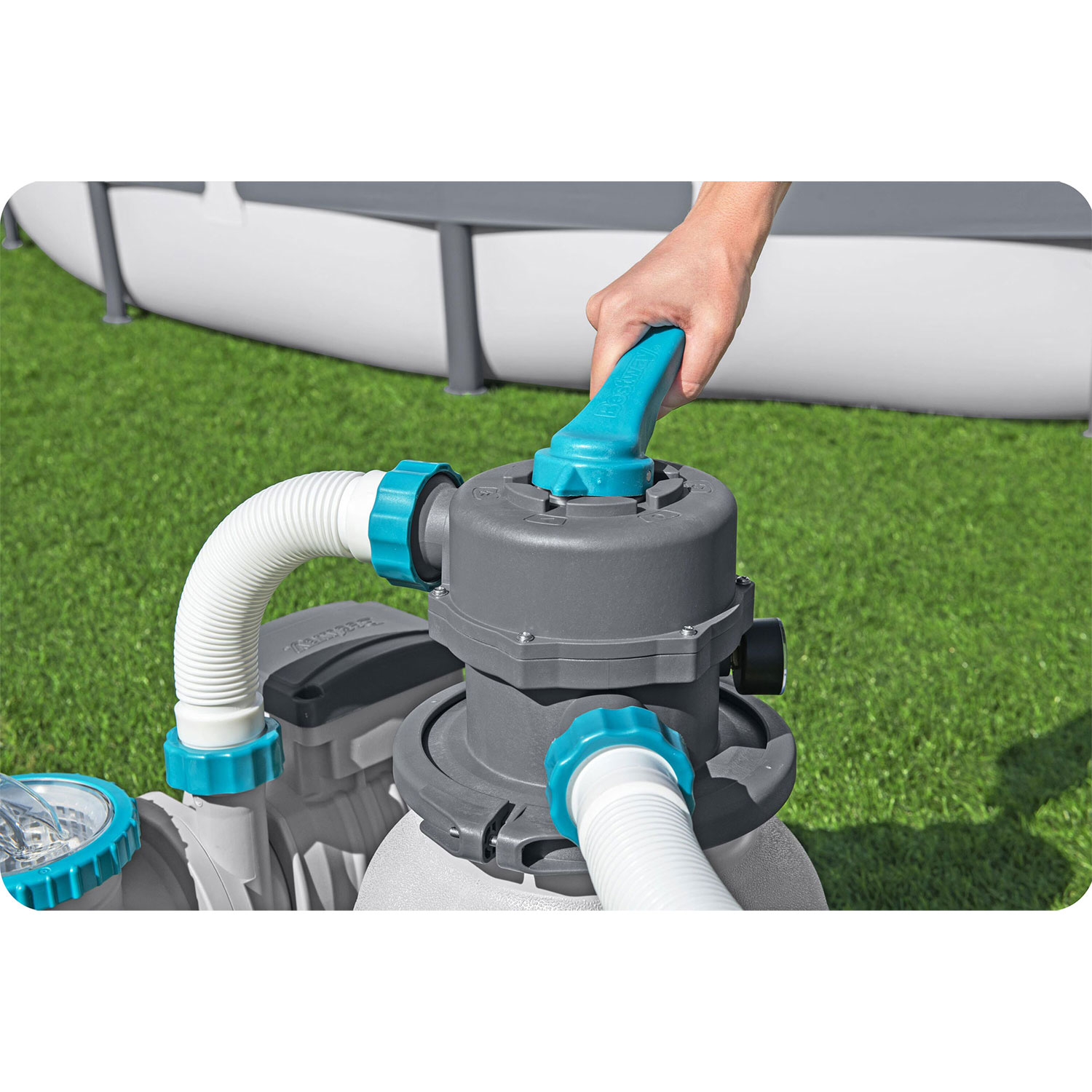 Ręka obsługuje filtr basenowy. Filtr jest szary z niebieskimi akcentami i białymi wężami. Basen jest w tle na zielonej trawie.