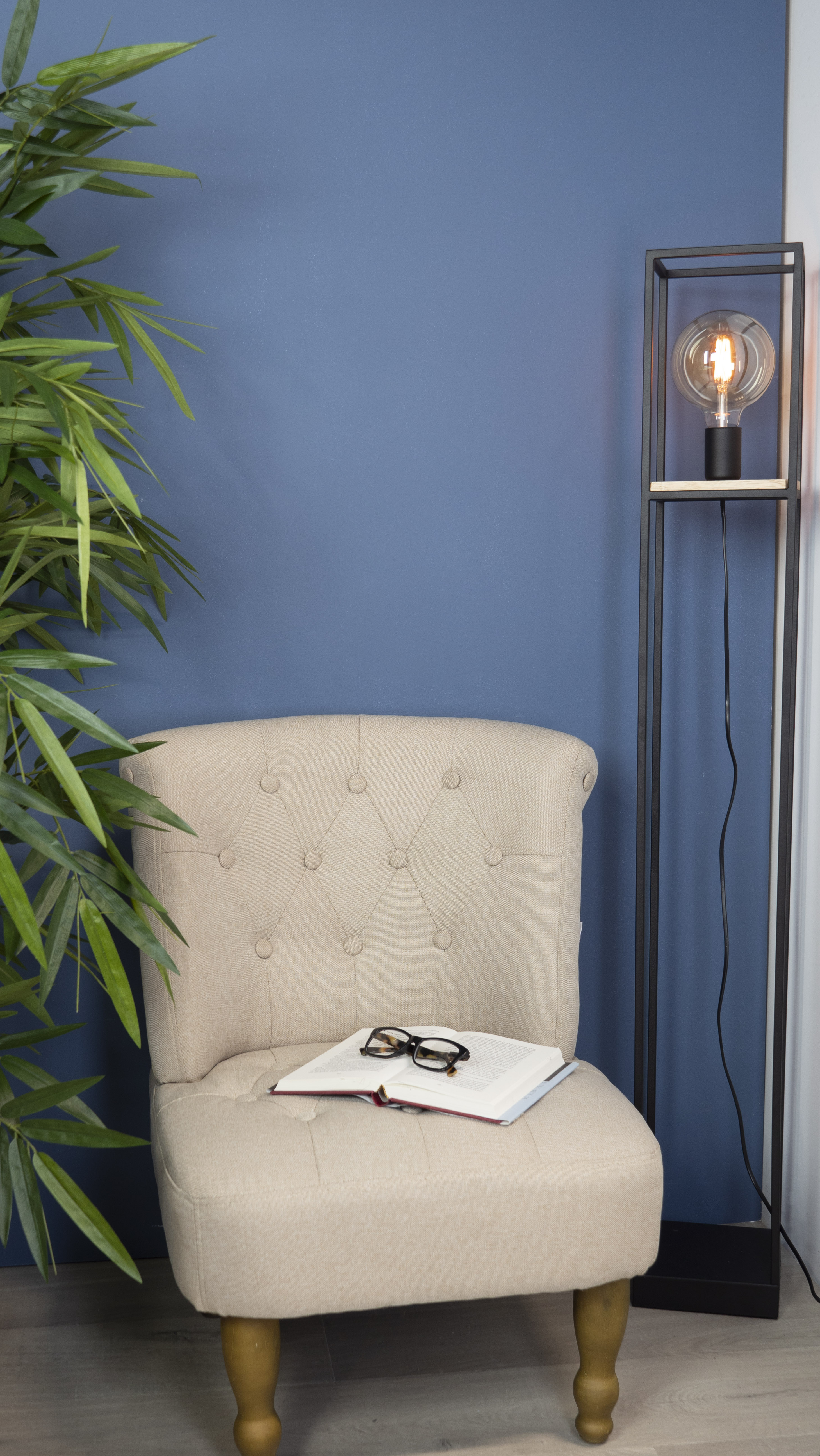Un fauteuil beige avec un livre et des lunettes est devant un mur bleu. Une lampe allumée et une plante verte sont également présentes.