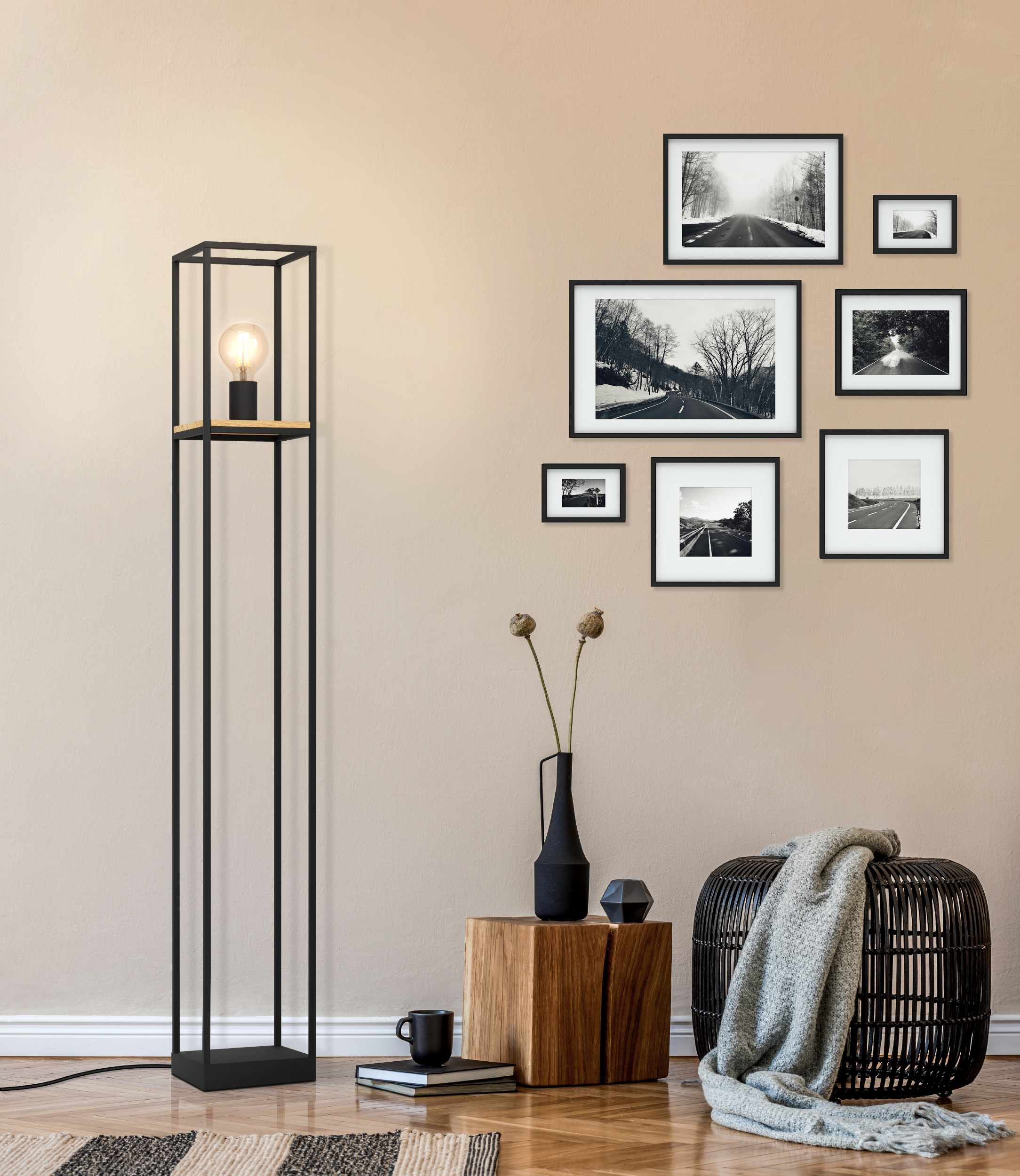 Un lampadaire noir, des tableaux, une table en bois avec décor, un tabouret tissé avec une couverture et un tapis.