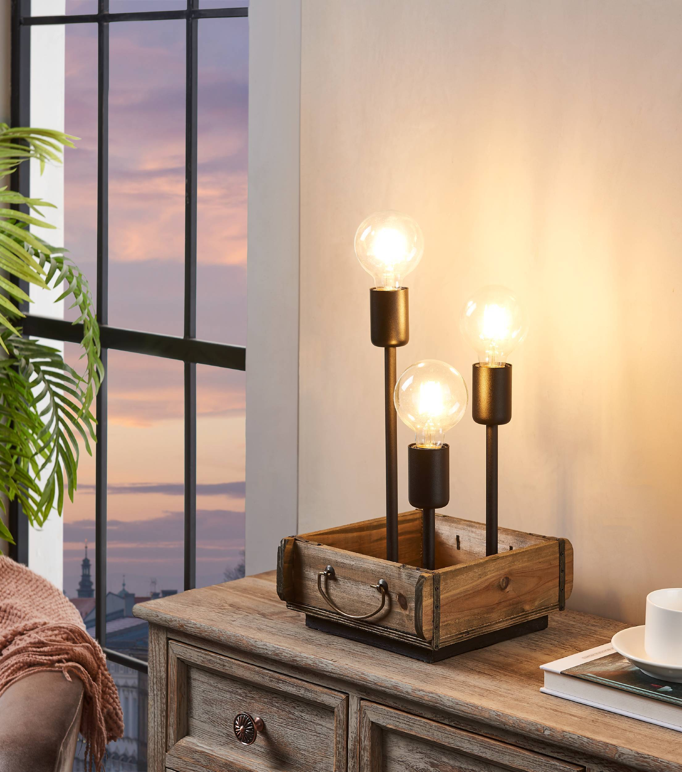 Eine Lampe mit drei Glühbirnen in einer Holzkiste steht auf einem Holztisch. Ein Fenster zeigt einen Sonnenuntergang.