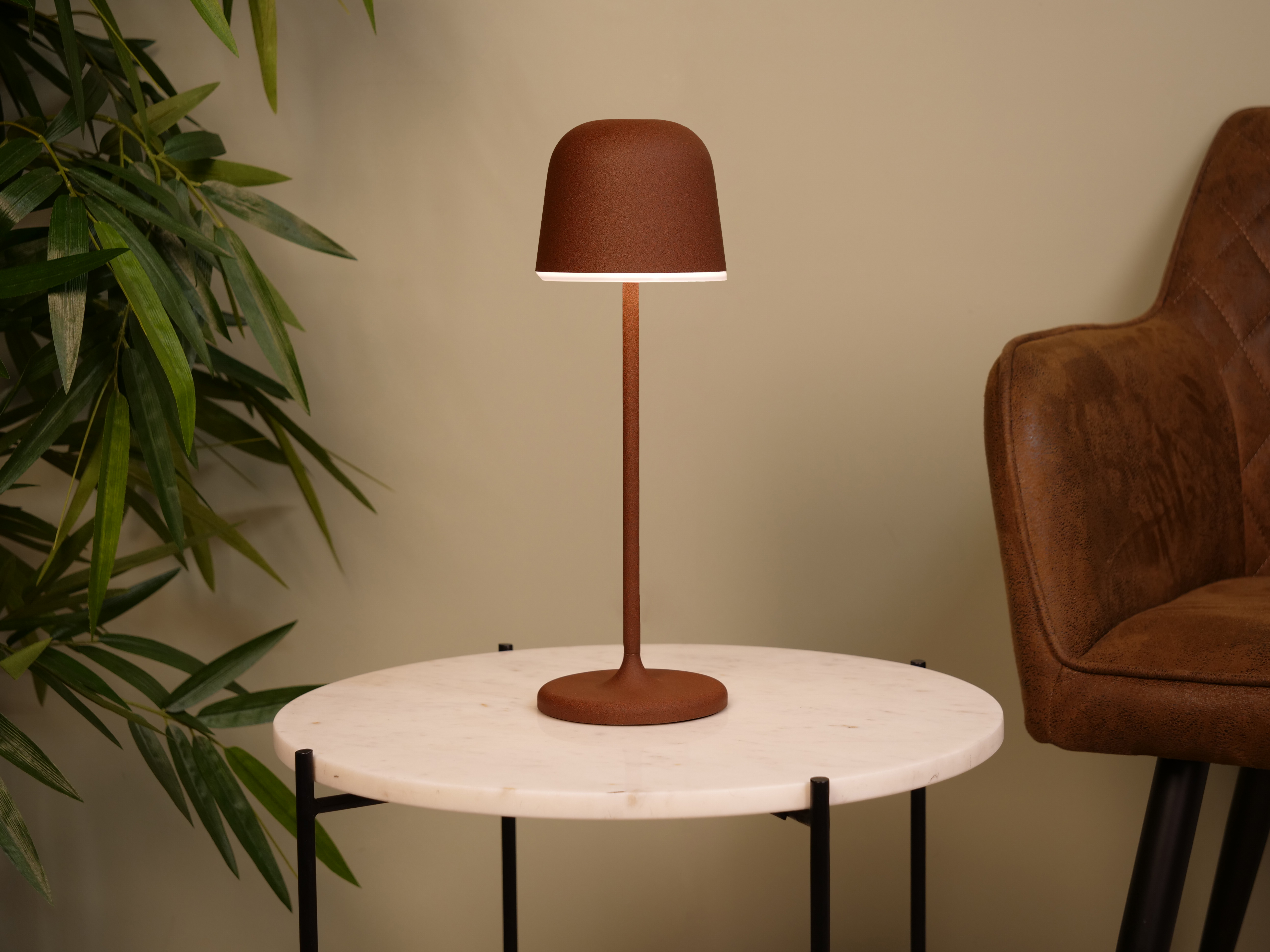 Eine braune Lampe auf einem Marmortisch, ein Lederstuhl und grüne Blätter vor beigefarbener Wand.