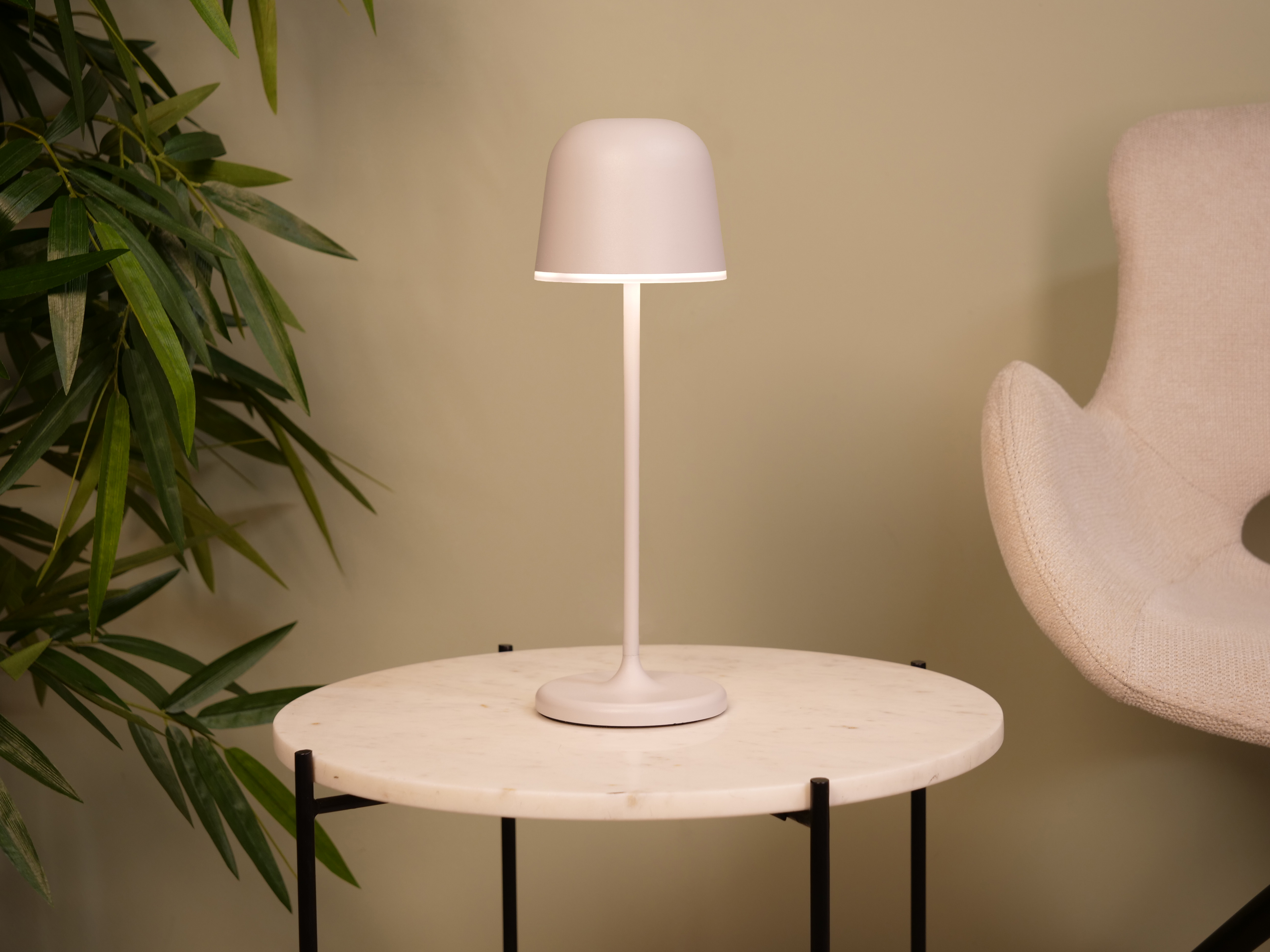 Eine kleine Lampe auf einem runden Tisch. Ein Stuhl und eine grüne Pflanze sind im Hintergrund.