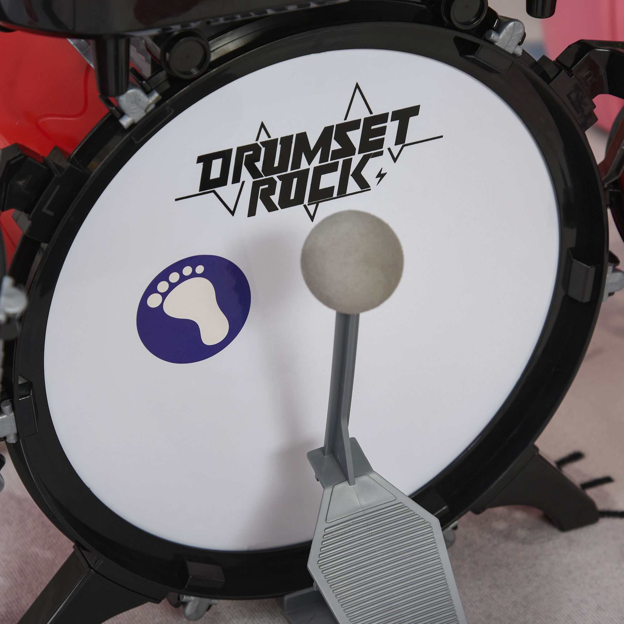 Primer plano de un tambor de juguete. El tambor tiene una superficie blanca con 'DRUMSET ROCK' escrito. Se ve un pedal y una baqueta.