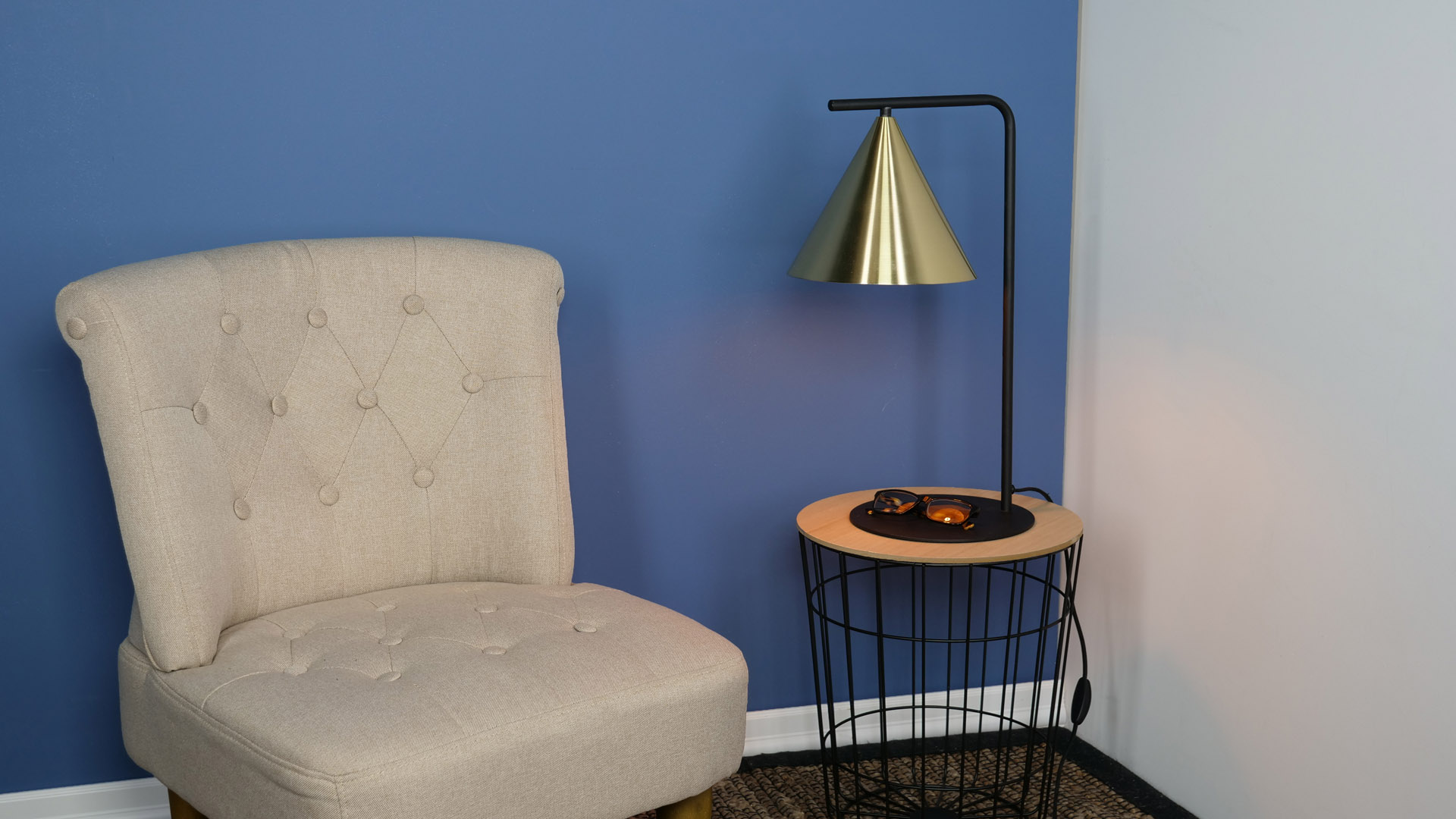 Ein beigefarbener Sessel, eine goldene Lampe und ein kleiner Beistelltisch mit Gläsern. Der Hintergrund sind blaue und weiße Wände.